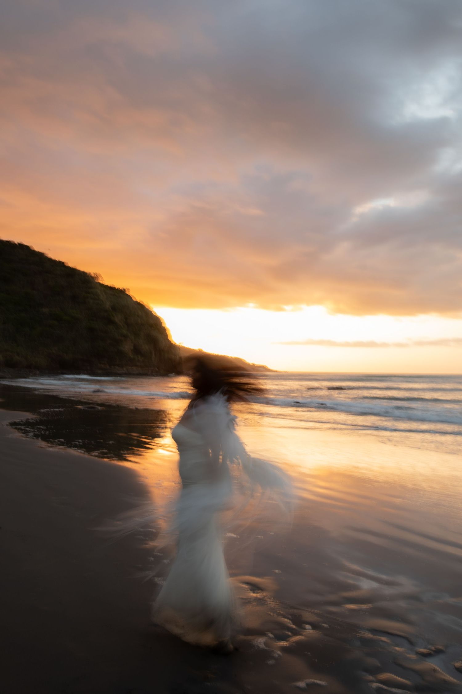 A blurry white figure moves across a beach at sunset creating a ghostly long exposure effect against the ocean waves.