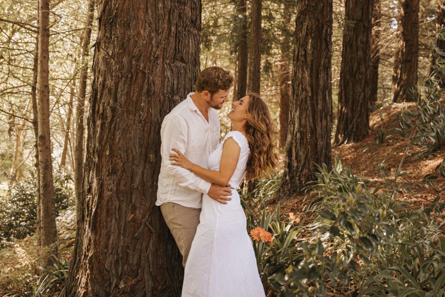 Romantic couple in white outfits share intimate moments during a forest photoshoot among tall trees.