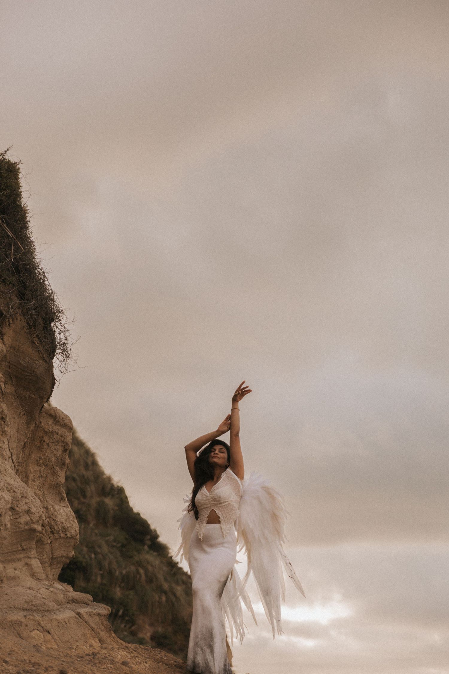 A figure in flowing white fabric dances gracefully against a dramatic cloudy sky and cliffside backdrop.