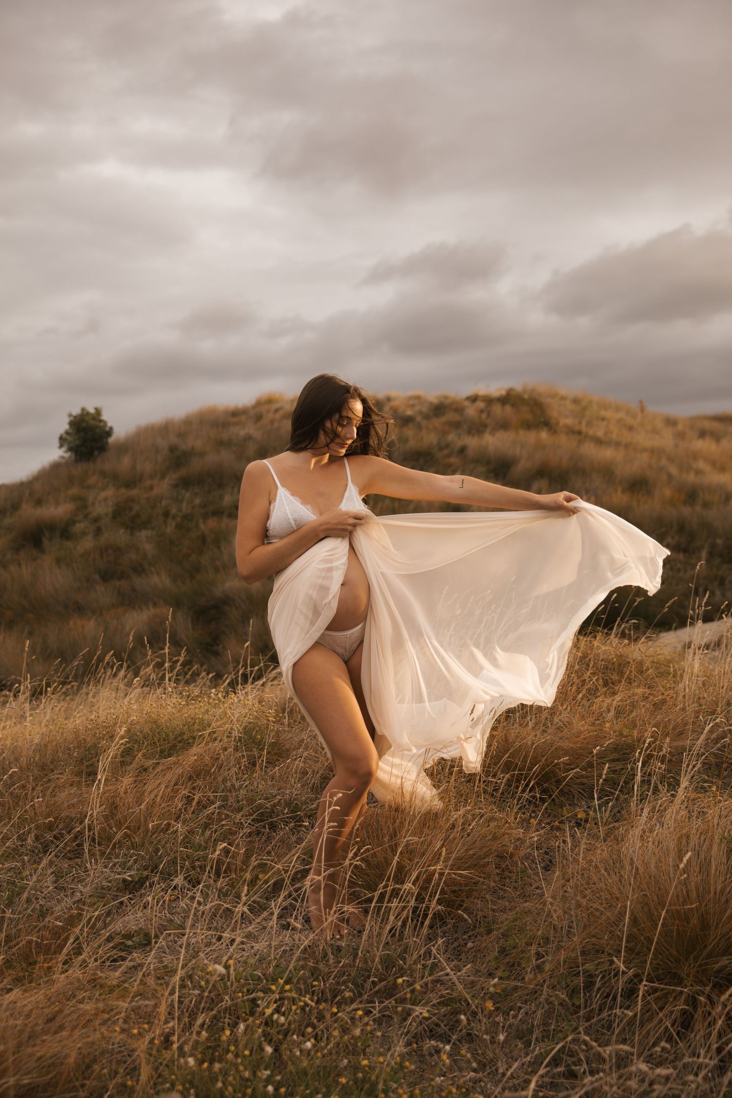Dreamy maternity portrait with flowing dress against golden hillside landscape.