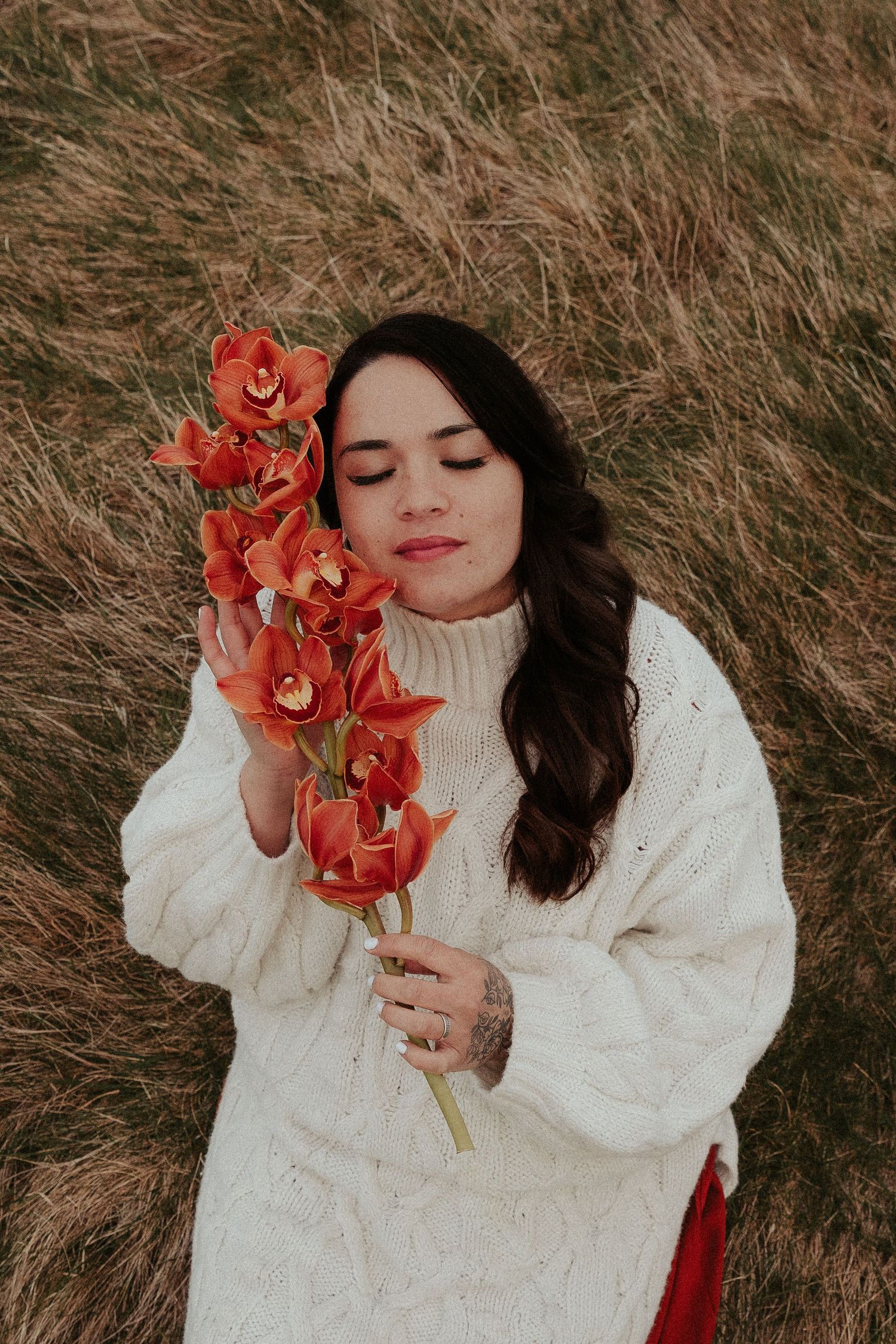 Person in white sweater poses with vibrant red orchids against dried grass background.