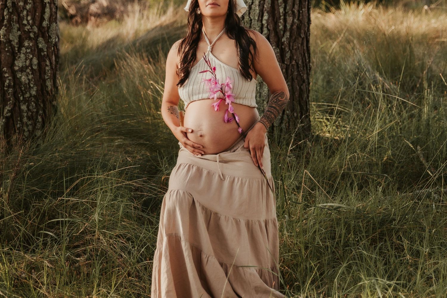 A maternity photoshoot in a sunlit field shows someone wearing a flowing beige dress and cradling their baby bump.