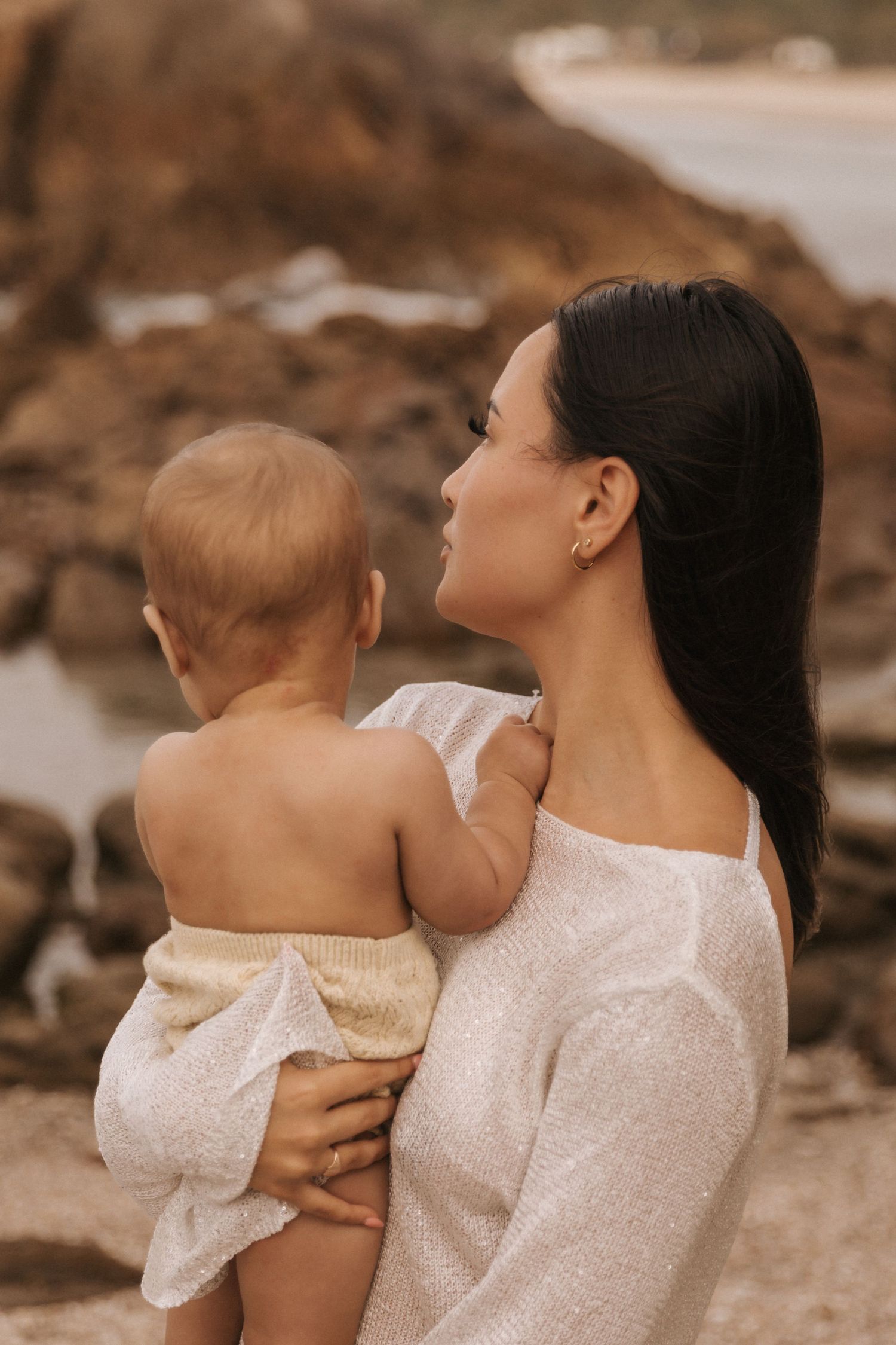 Mother and baby share joyful moment against rocky coastal backdrop at sunset.