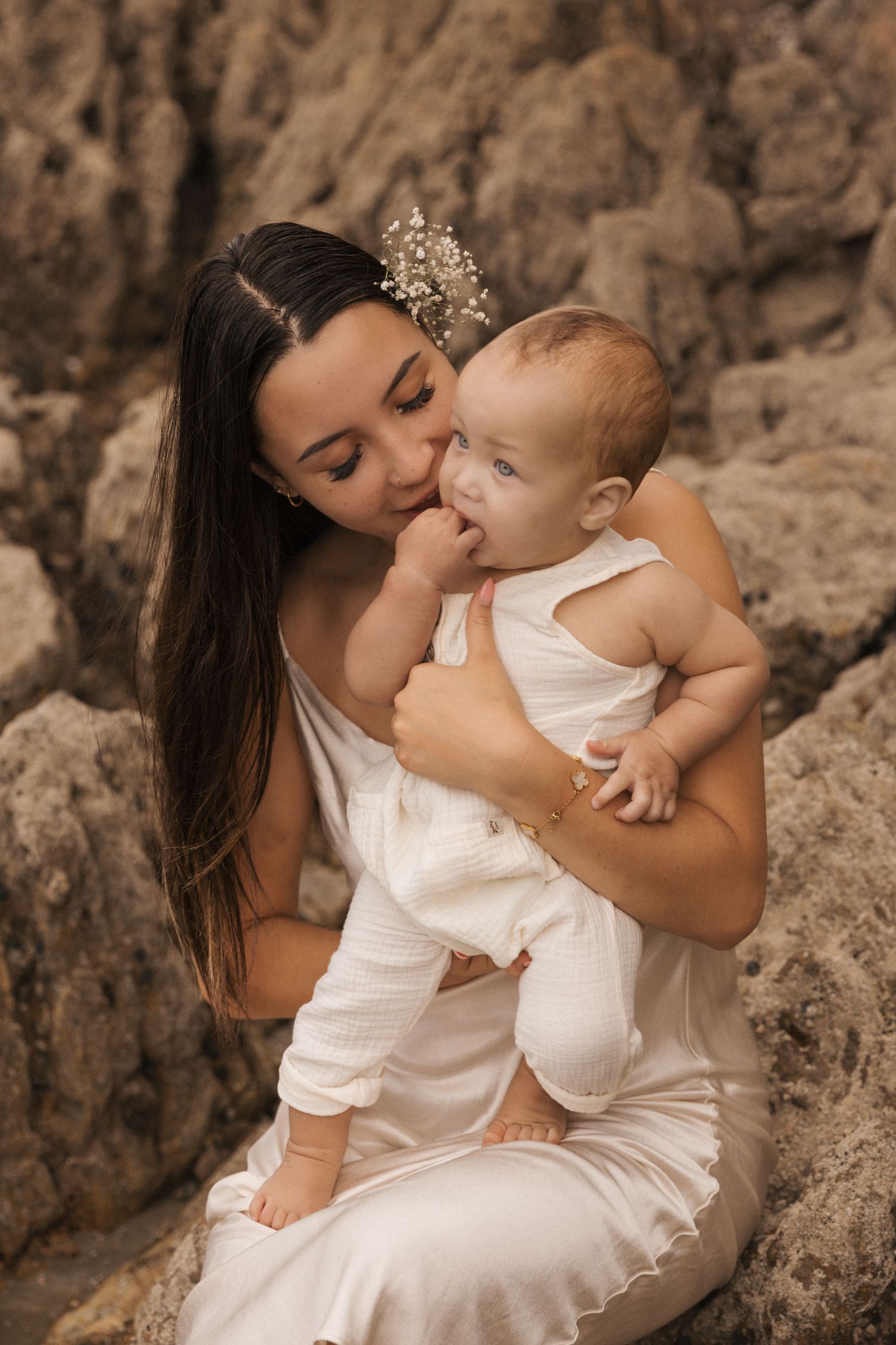 Intimate mother-child portrait against textured rock wall in white clothing.