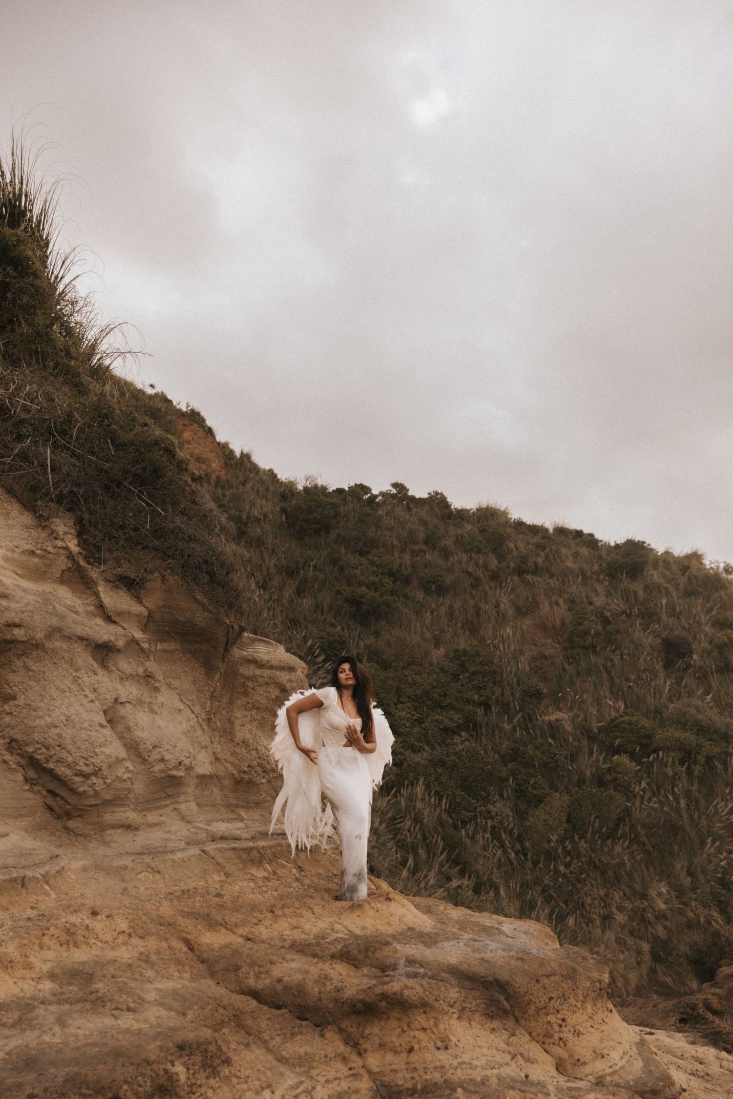 A couple in white attire poses together on a scenic cliffside overlooking a natural landscape at sunset.