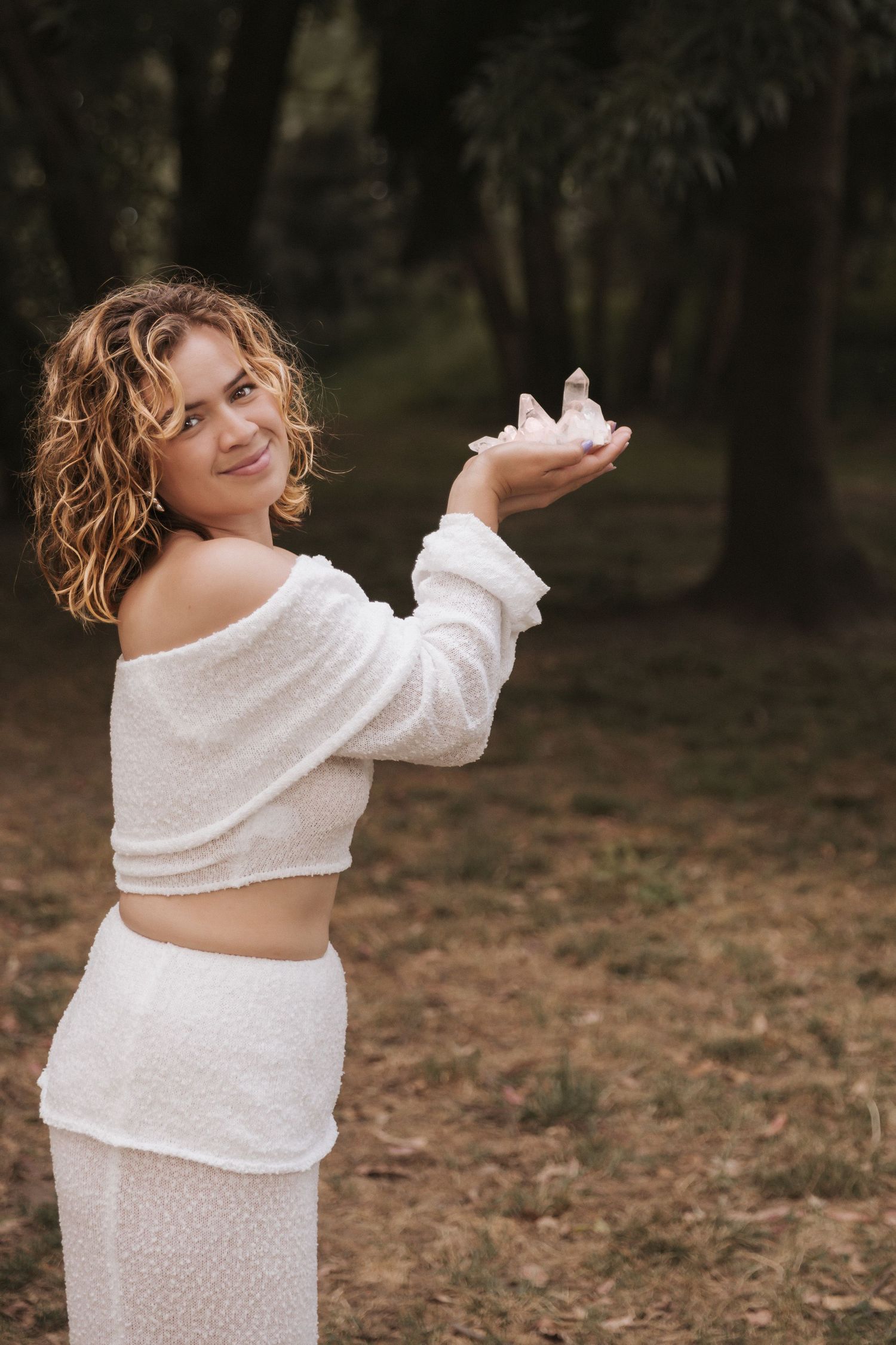 Woman in white crop top and skirt poses playfully in forest setting.