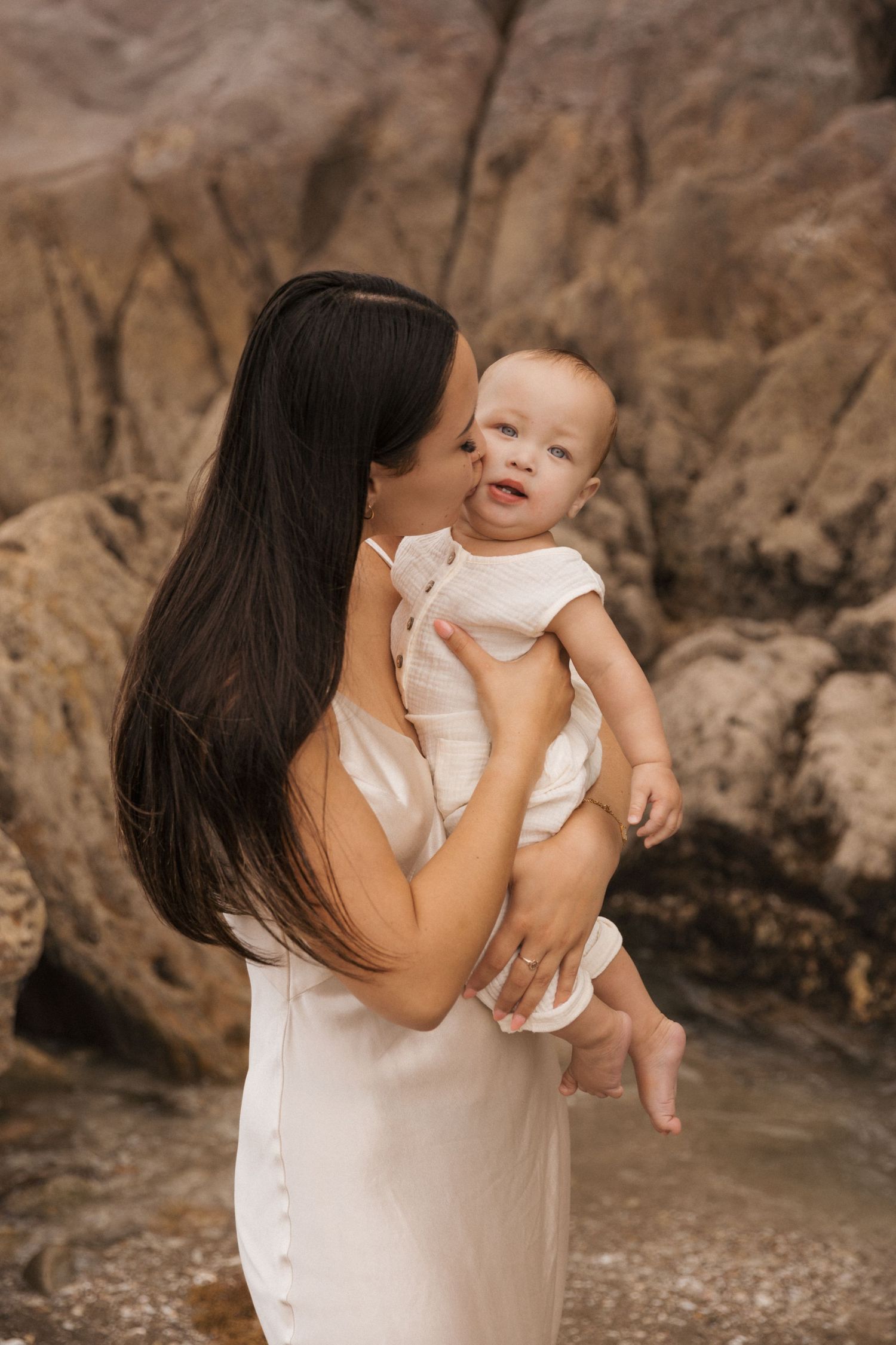 Mother holding baby in white dress against rocky beach background.