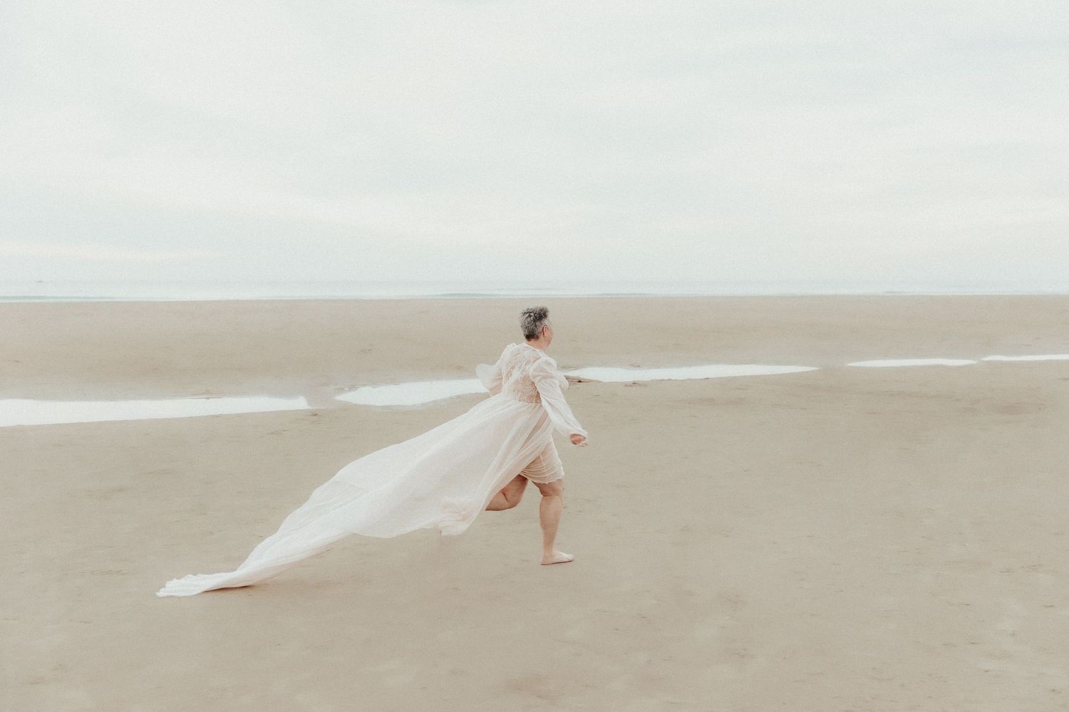 Figure running along empty beach with flowing white fabric catching the ocean breeze.
