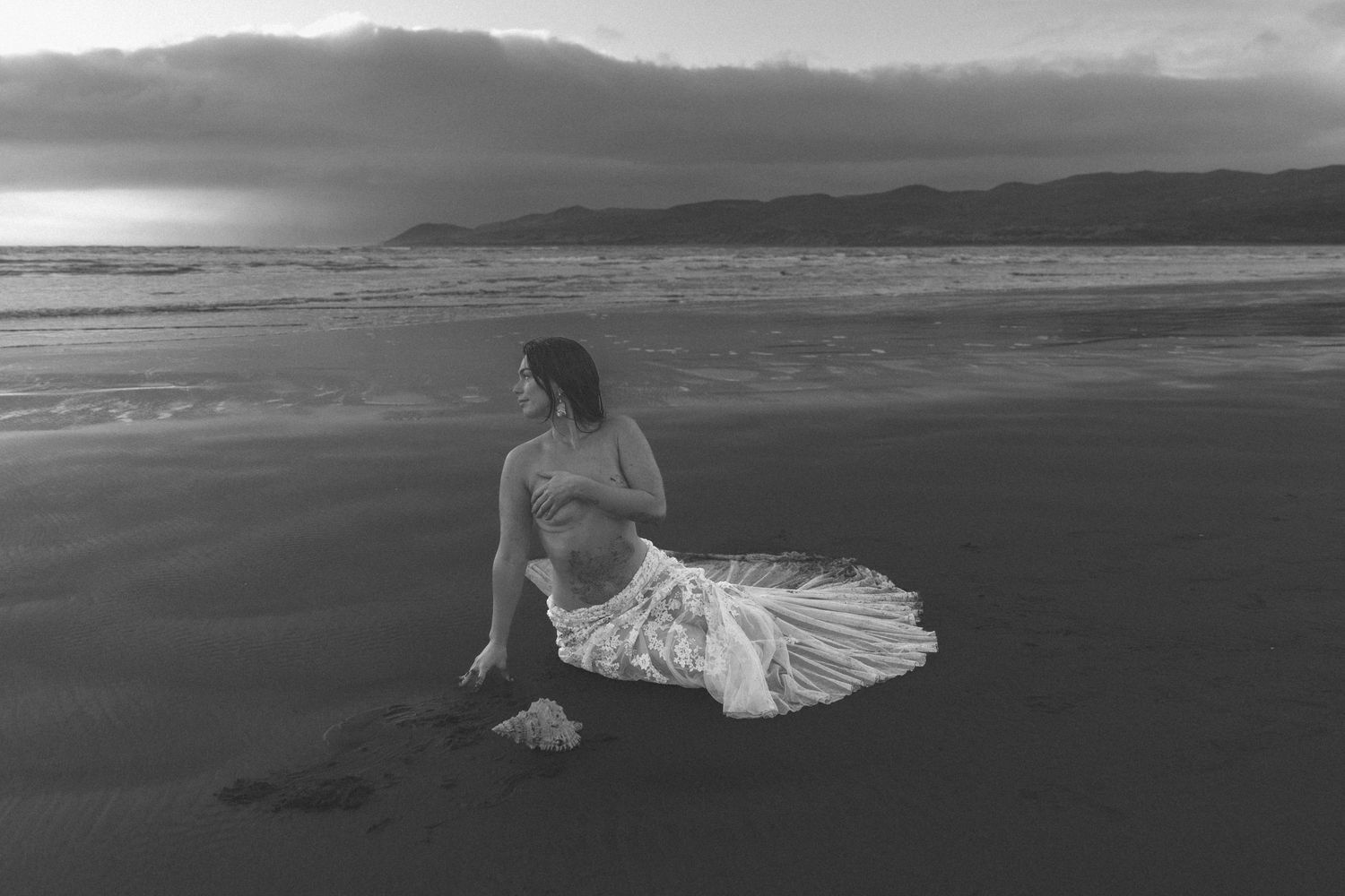 A moody black and white beach scene with mountains in the distance and flowing white dress on dark sand.