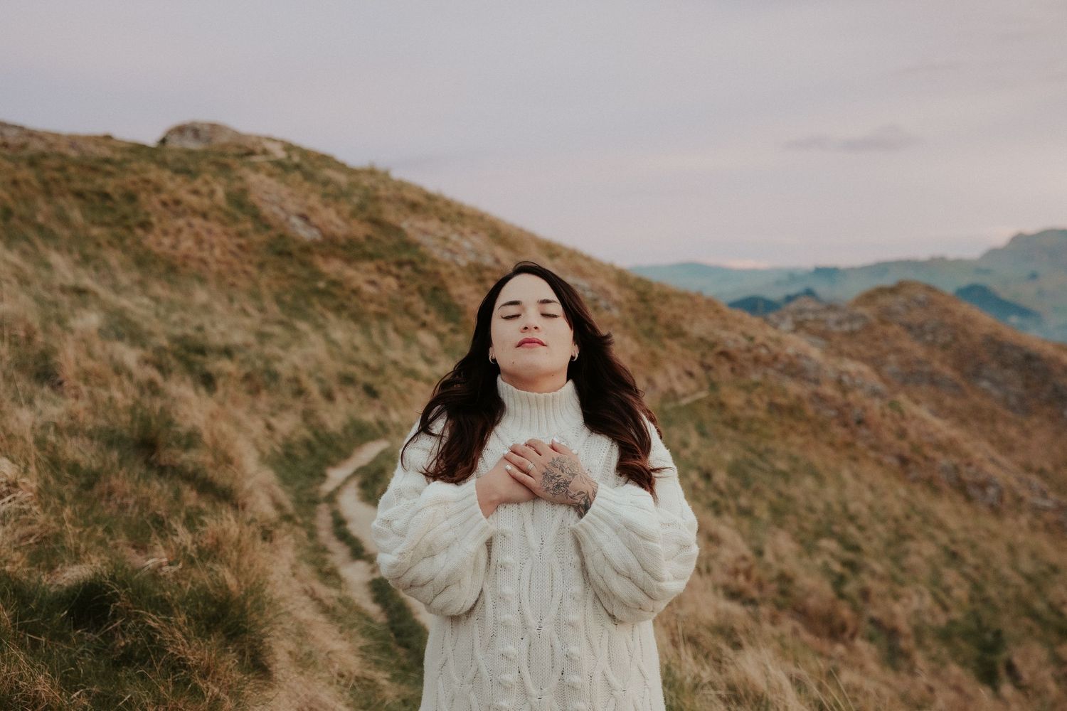 Person in white dress poses expressively against rolling hills and cloudy sky.