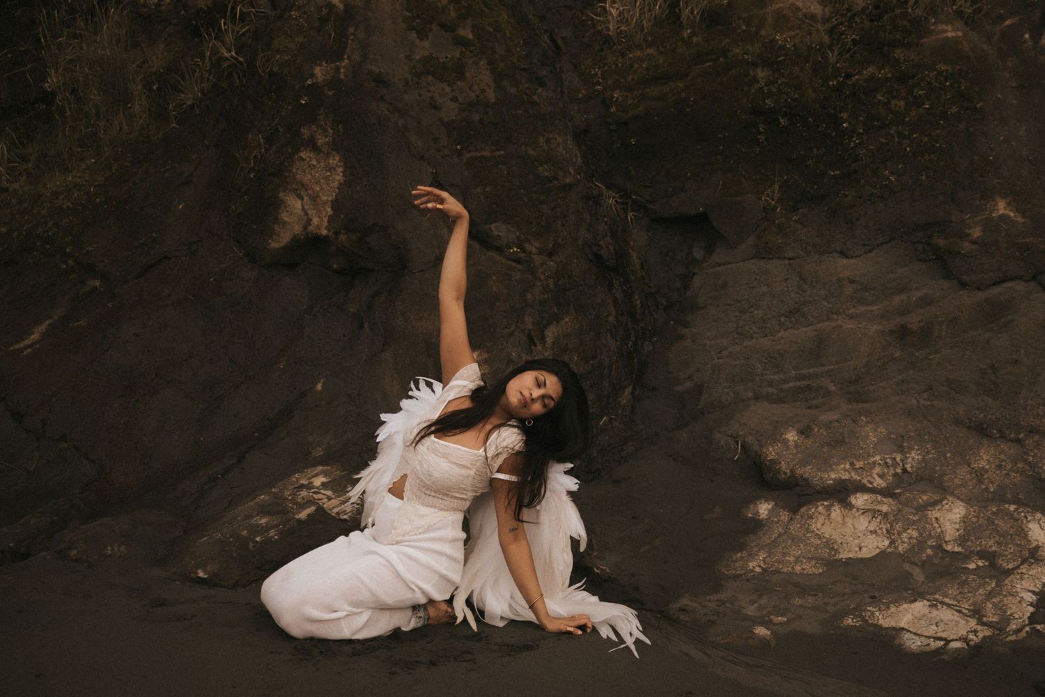 A couple in white wedding attire poses dramatically on dark rocks during a romantic photo session.