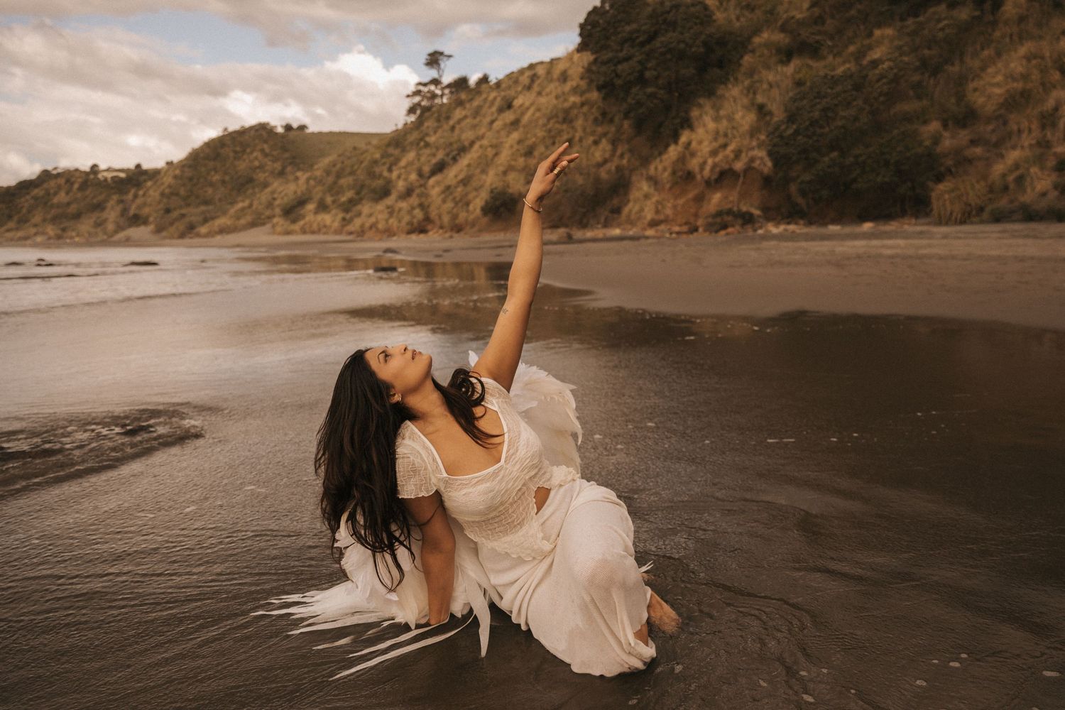 A dancer in a flowing white dress performs an elegant pose on a dramatic beach at sunset with cliffs in the background.