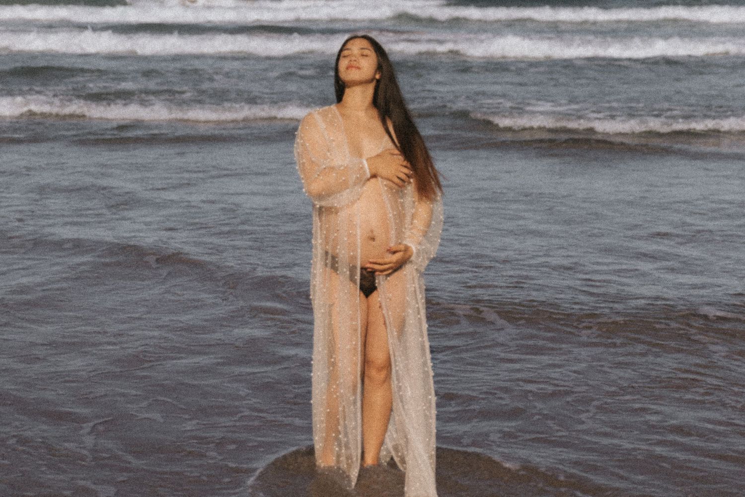 Beachside maternity session with flowing peach dress against ocean waves.