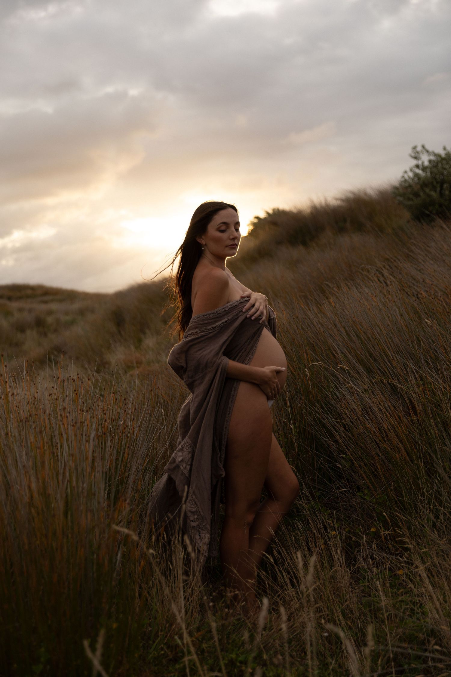 Artistic maternity silhouette against sunset sky with flowing fabric and tall grass.