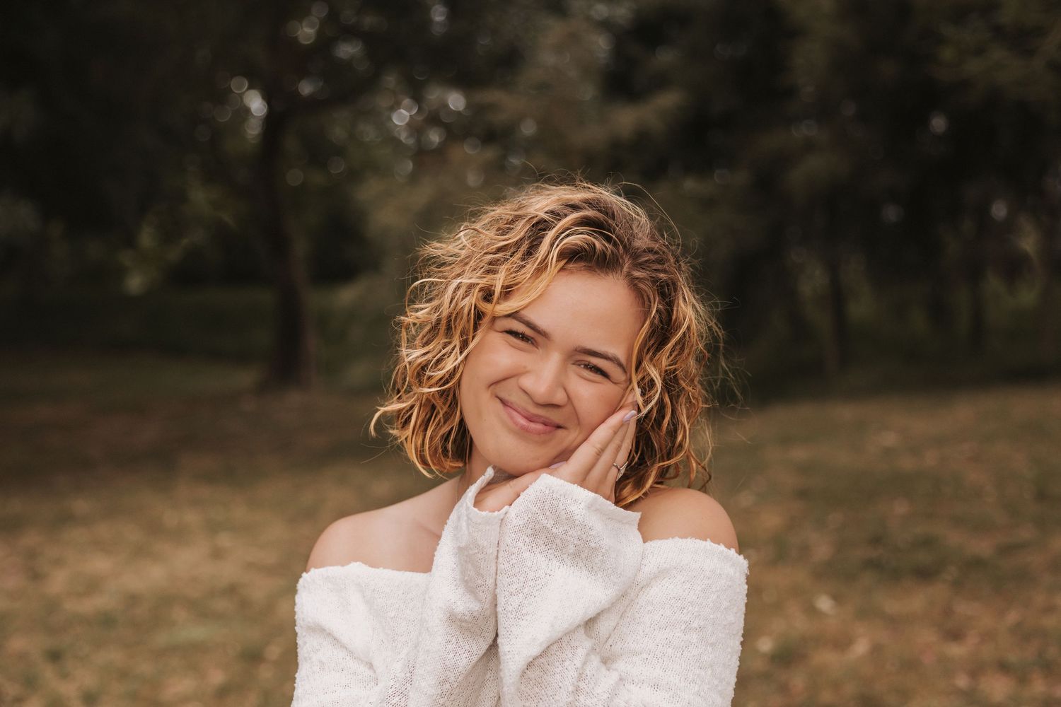Woman with curly hair in white sweater smiles warmly in natural outdoor setting.