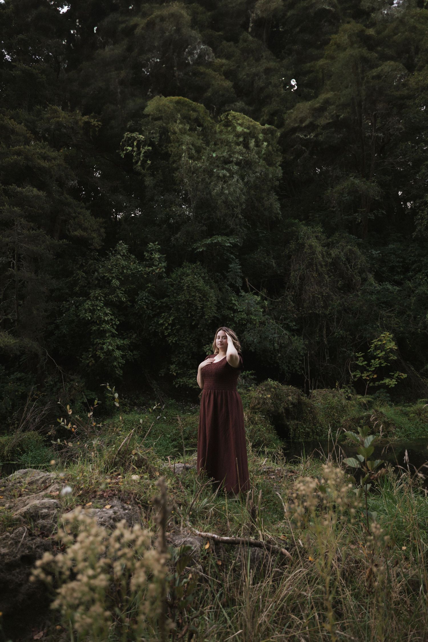 A moody forest portrait with a figure in a burgundy dress standing on rocky terrain.