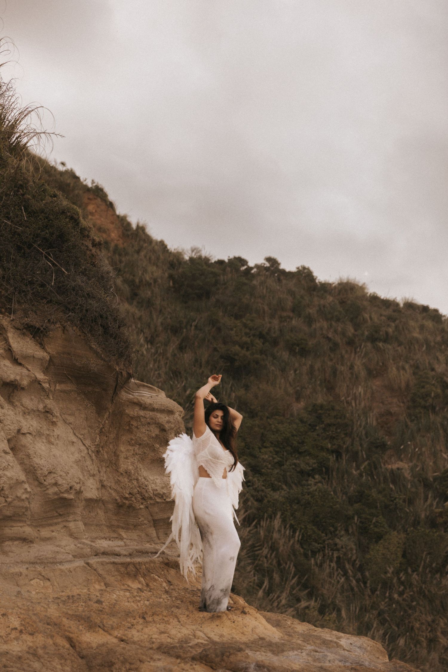 A couple in white attire poses gracefully on a rocky cliff edge against a moody cloudy sky and hillside backdrop.
