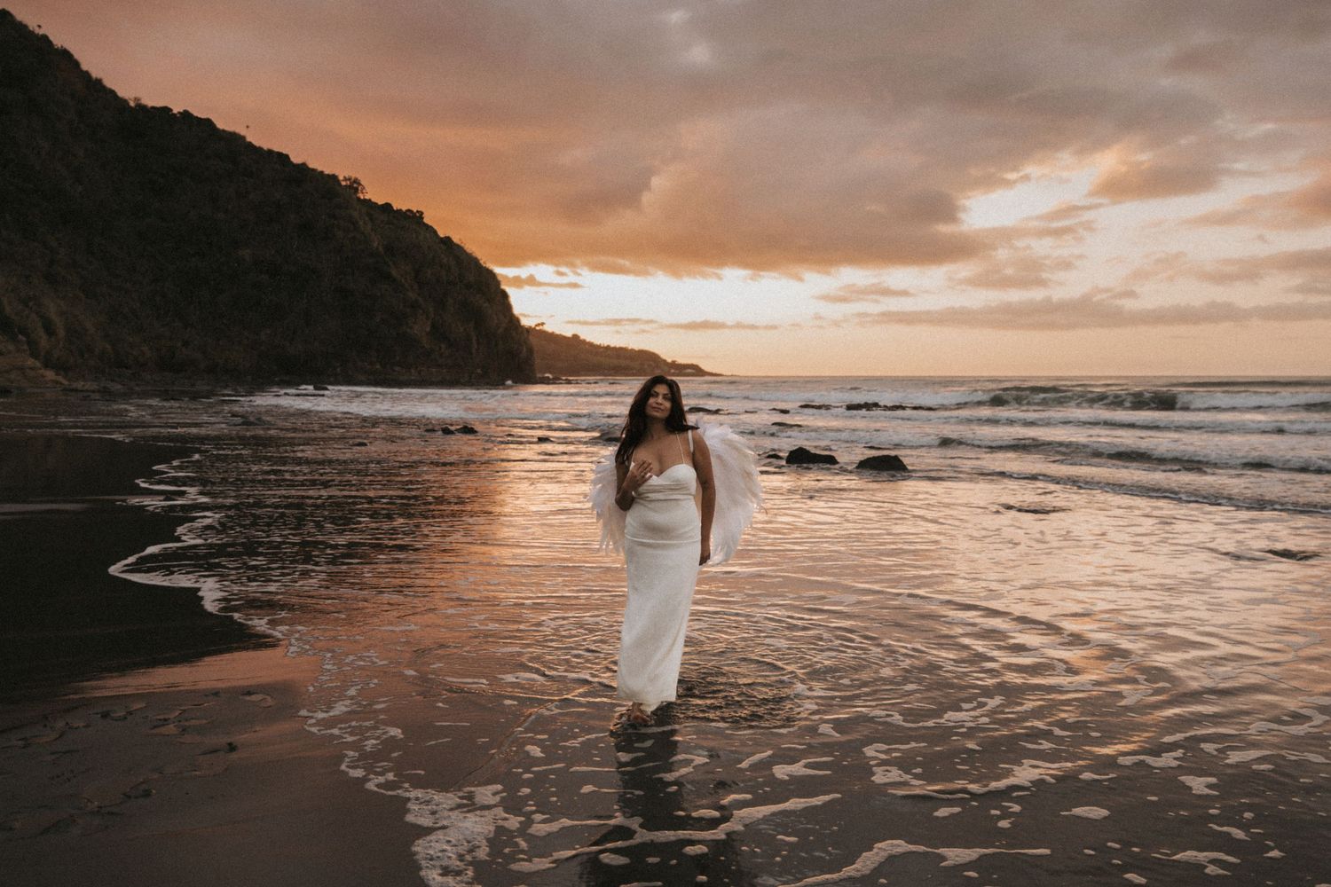 A person in a flowing white dress walks along a scenic beach at sunset with dramatic orange clouds in the sky.