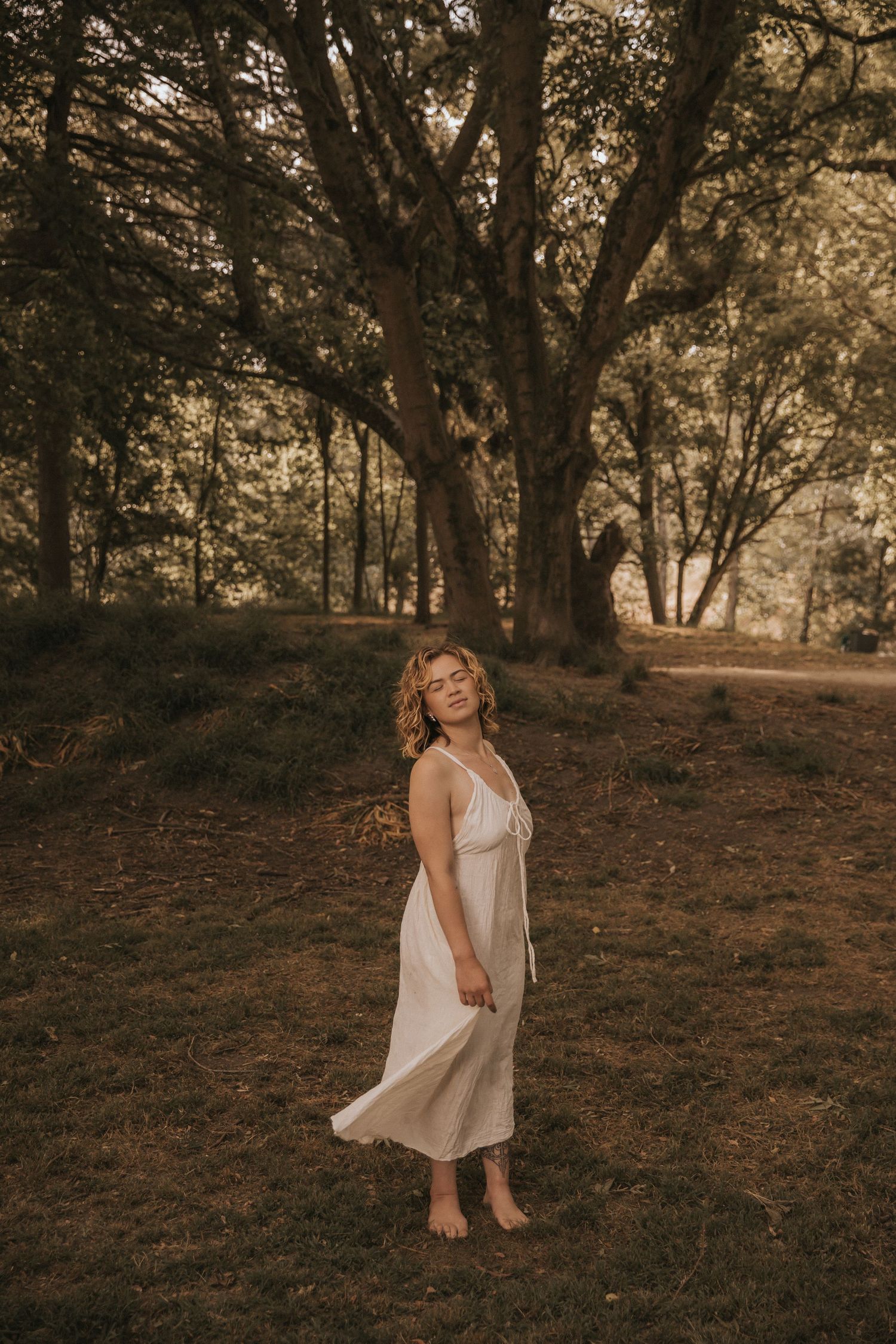 A ethereal portrait in a white dress standing barefoot among tall trees at sunset.