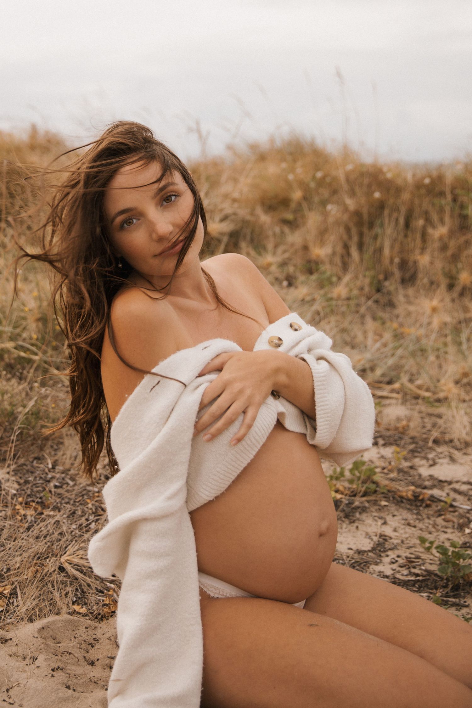Intimate maternity portrait in white wrap against coastal dunes at golden hour.
