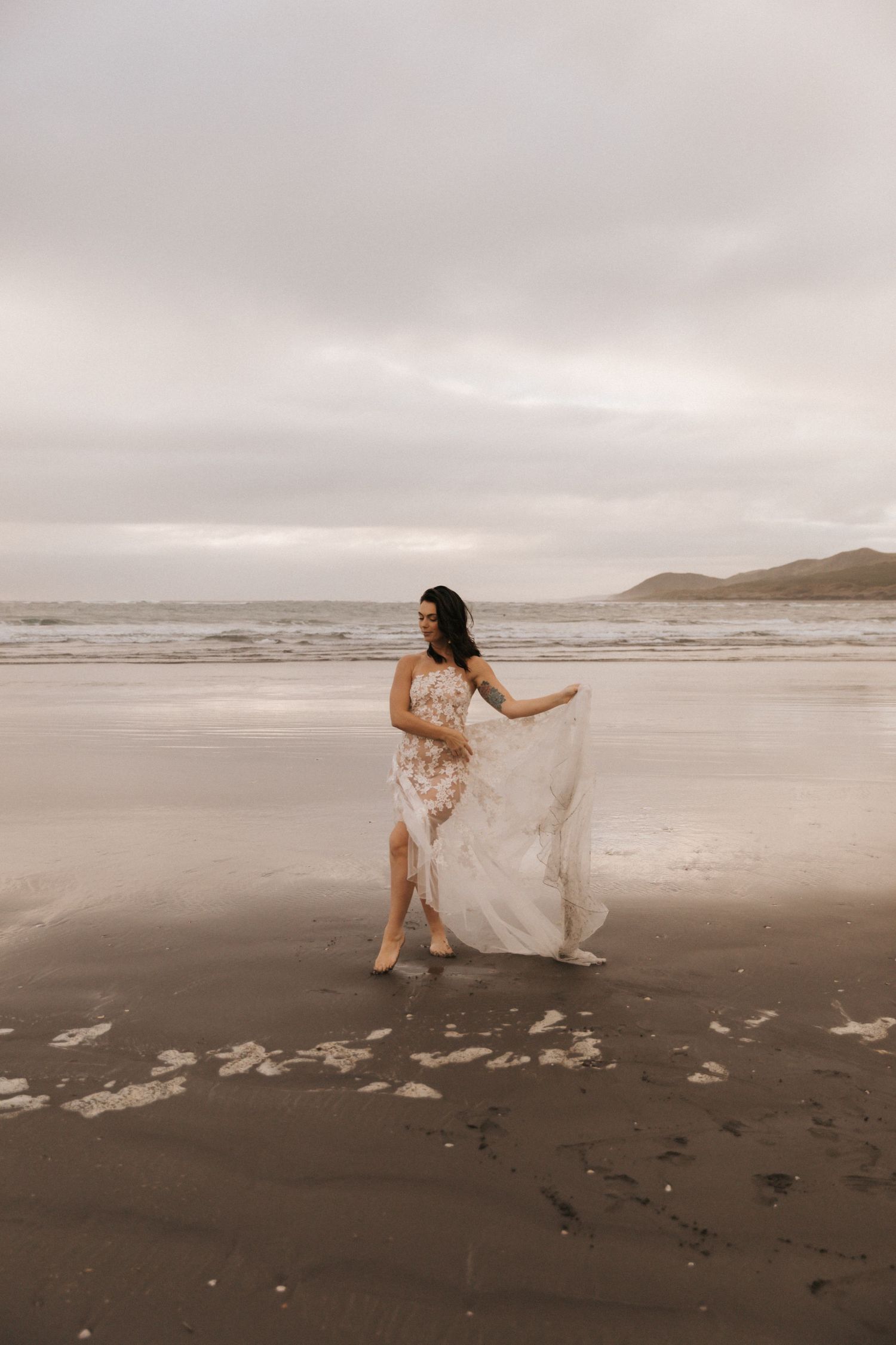 Artistic beach portrait with bride dancing in delicate lace gown at low tide.