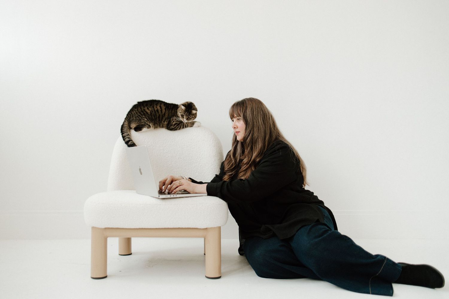 A person in dark clothing lies on a white floor while reaching for a small wooden stool.