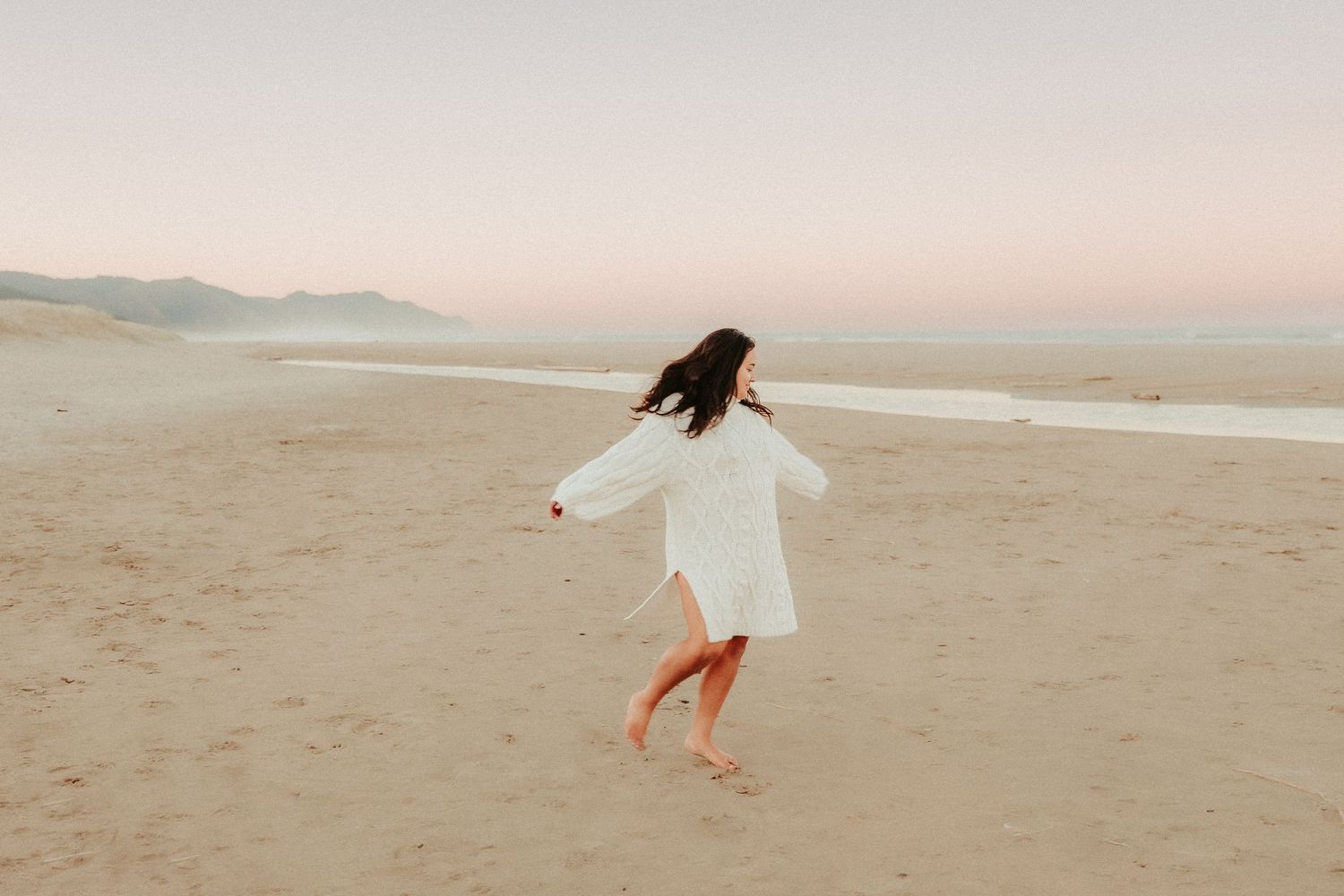 Woman in flowing white dress dancing on beach at sunset.