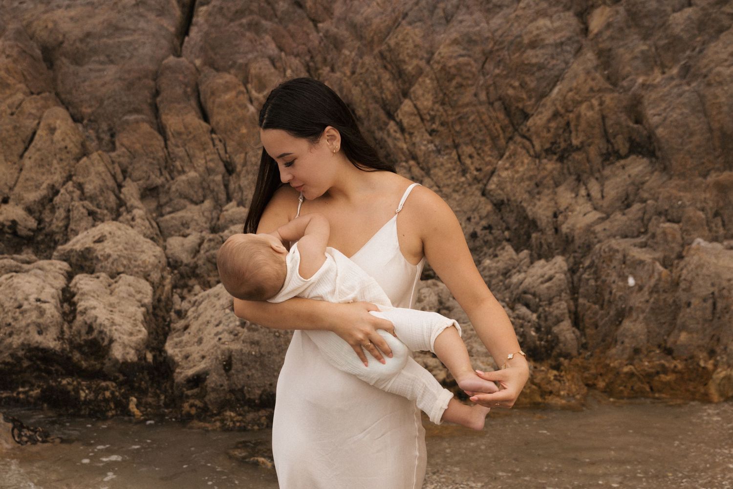 Mother and baby in white dresses posing by coastal rocks.