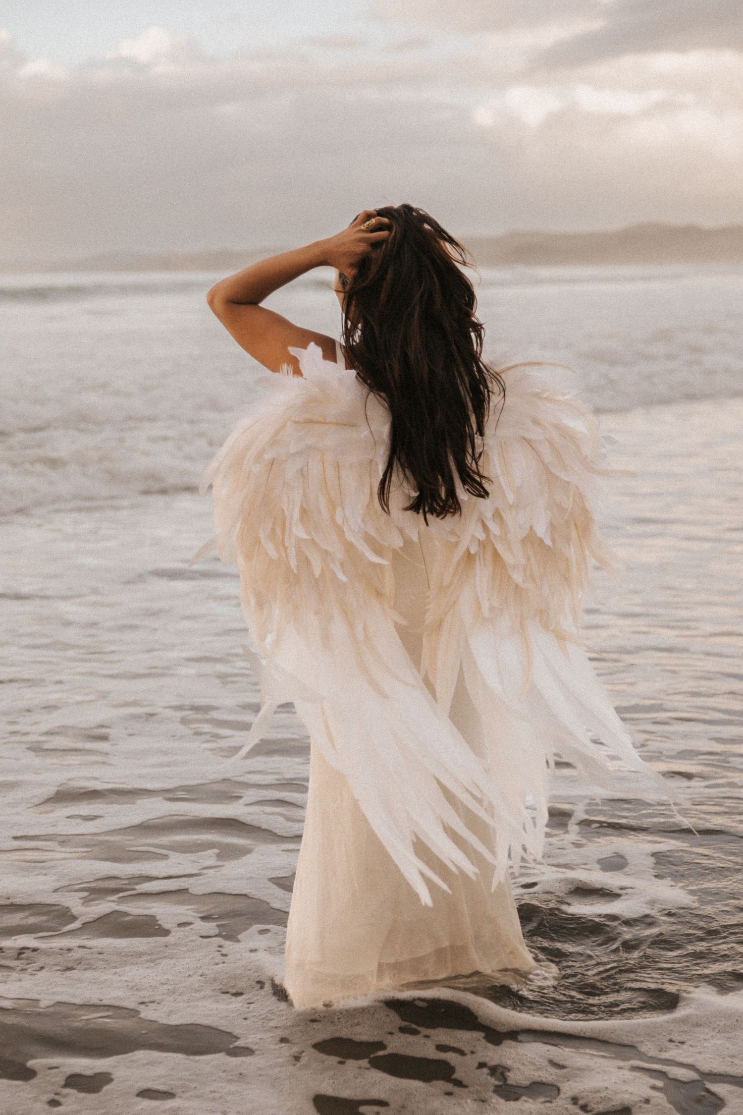 A person in a flowing white dress with feathered angel wings stands on a beach at sunset looking at the ocean.
