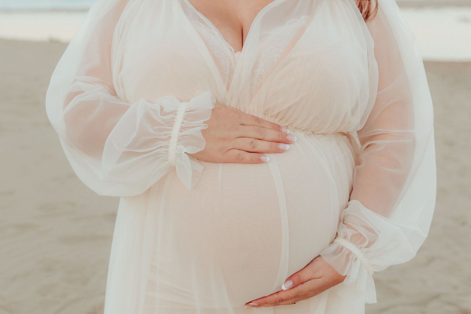 A woman in a flowing white dress holds their baby bump while standing on a sandy beach at sunset.