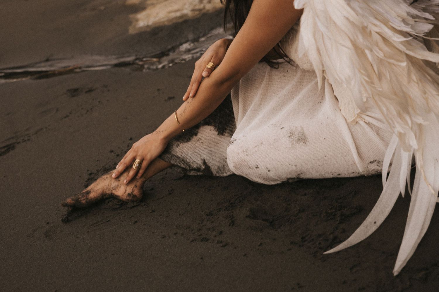 Hand wearing white feathered dress touches black sand on a moody beach in artistic wedding photo.