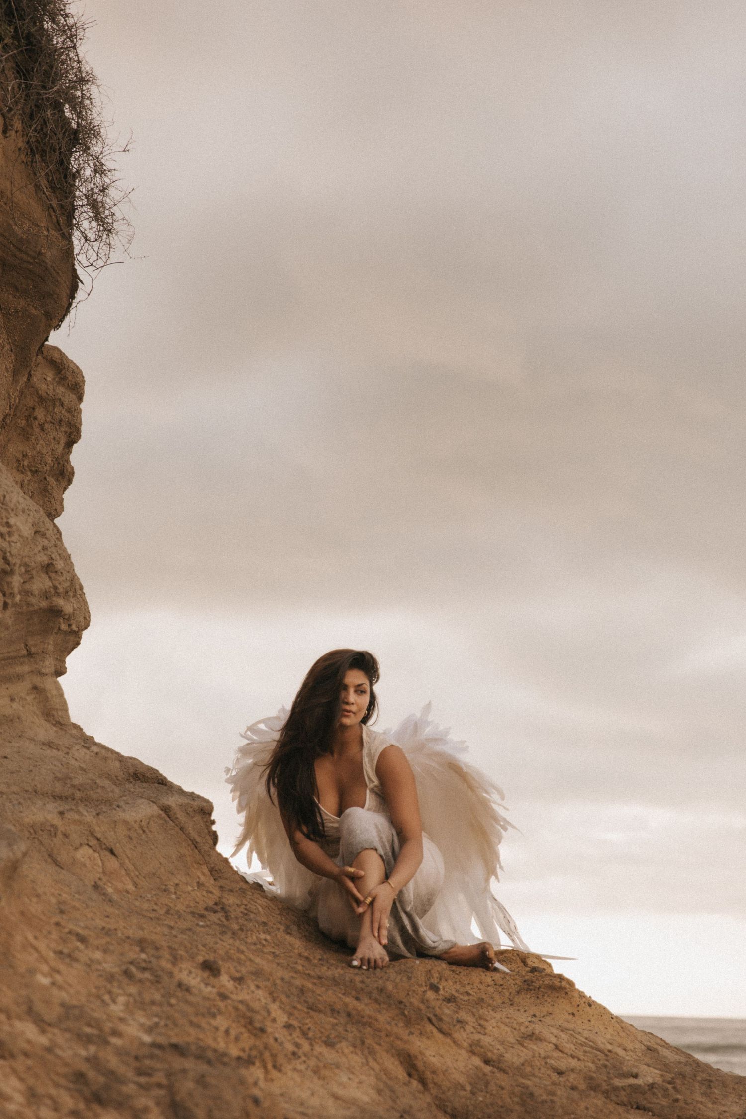 A figure wearing white angel wings poses dramatically on a rocky cliff against a moody cloudy sky.