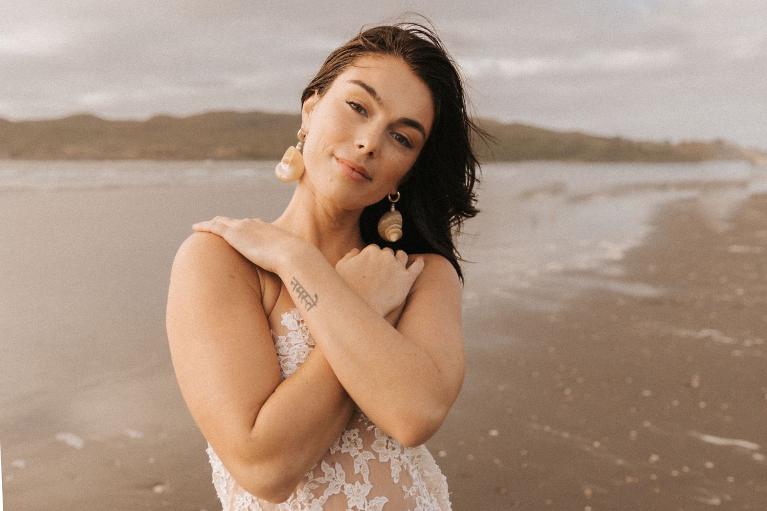 Woman in white lace dress poses expressively on beach.