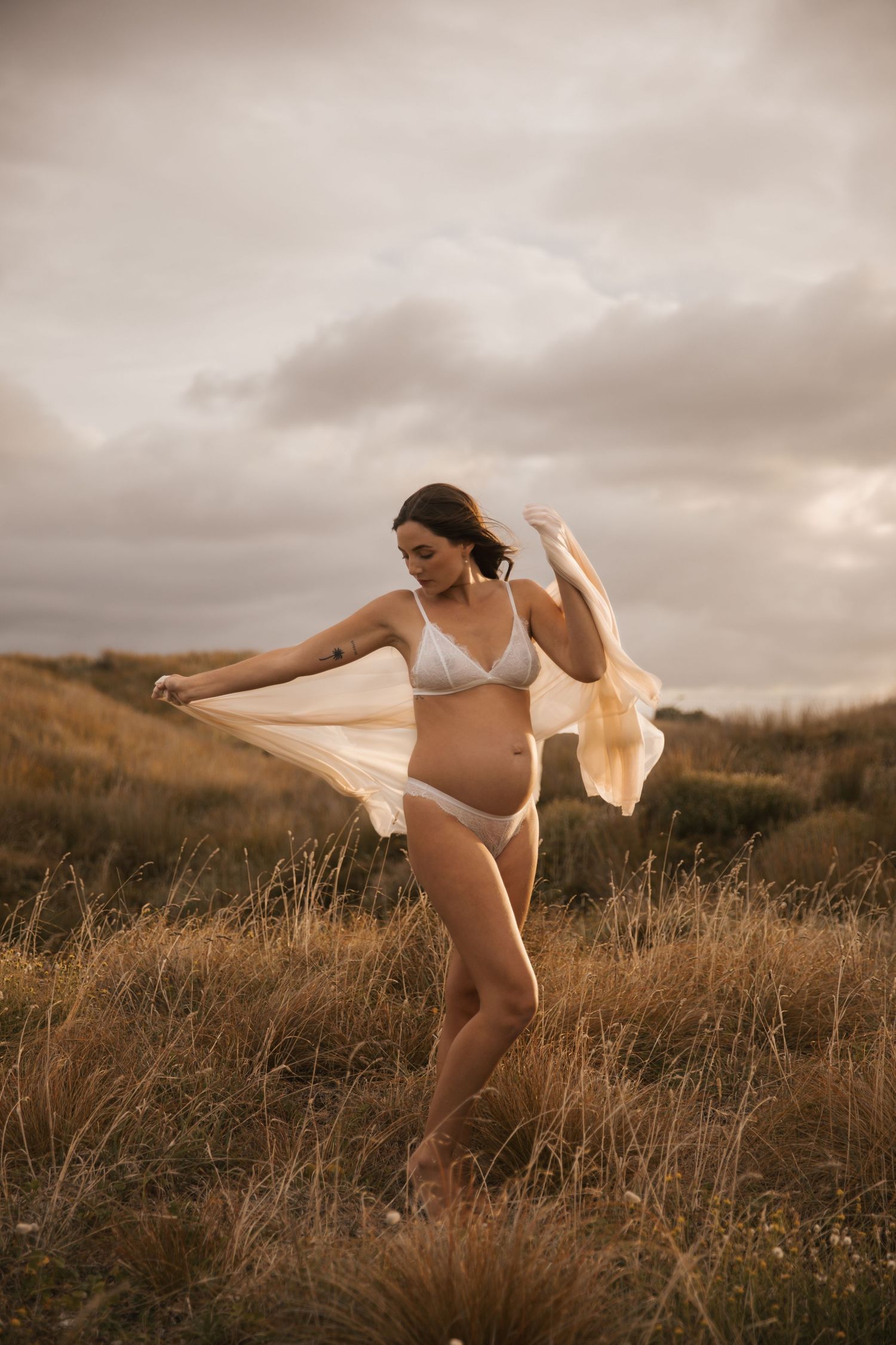 Artistic maternity photo with flowing fabric against moody sky and golden grass.