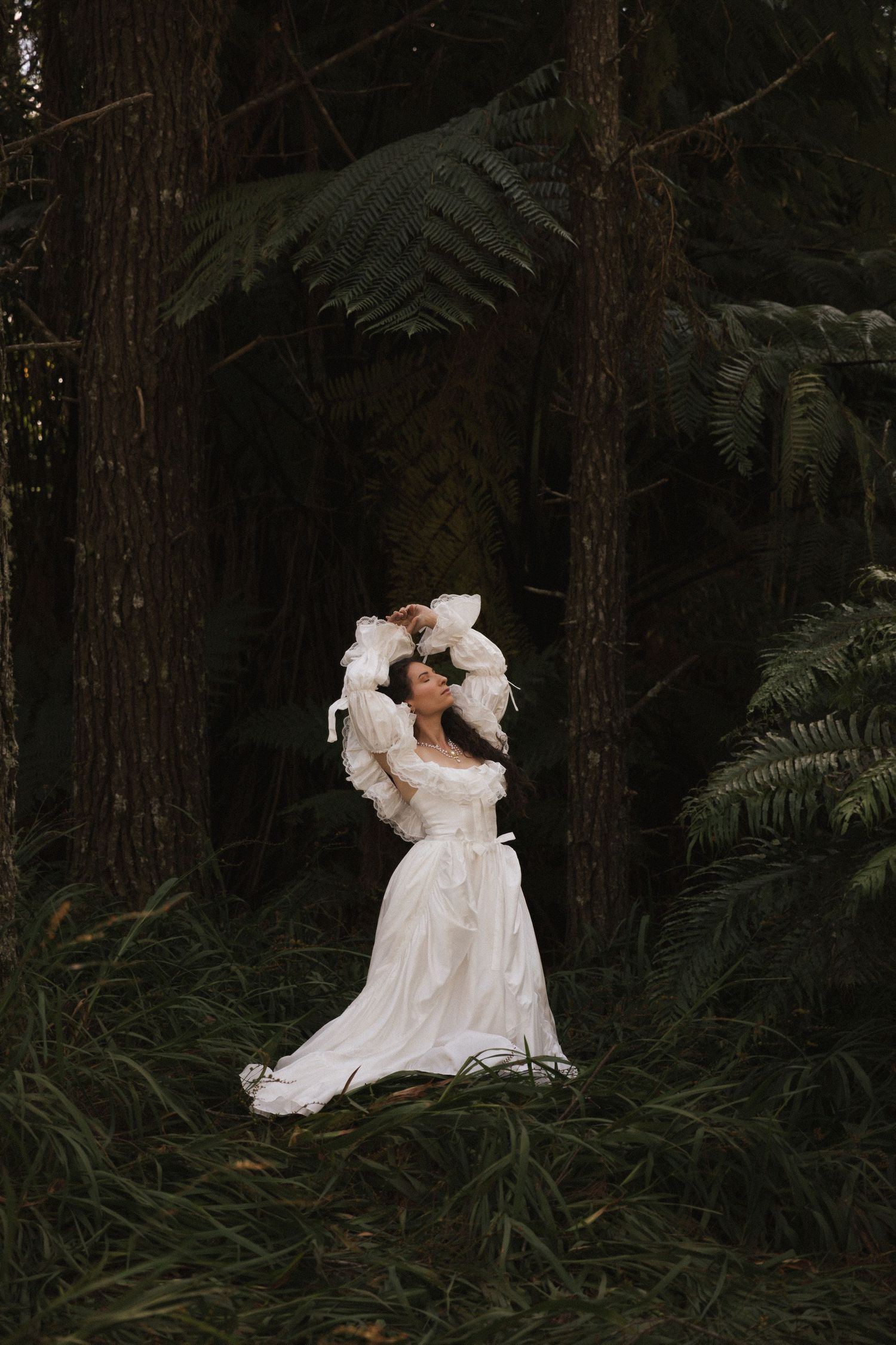 Woman in white vintage dress poses gracefully among tall ferns and trees in a lush, dark forest setting.