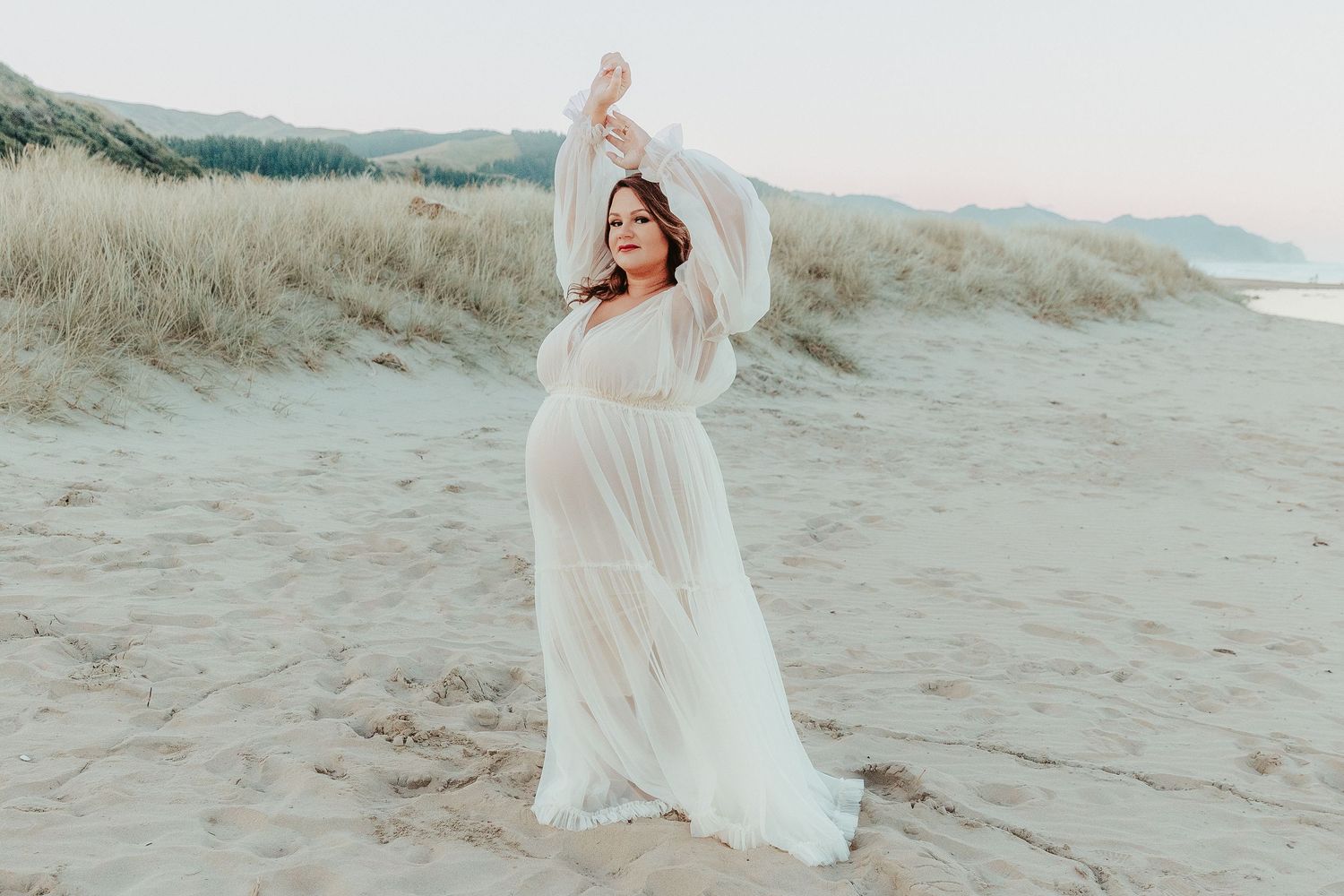 Ethereal maternity photo of a woman in flowing white dress dances gracefully on sandy beach at dusk.