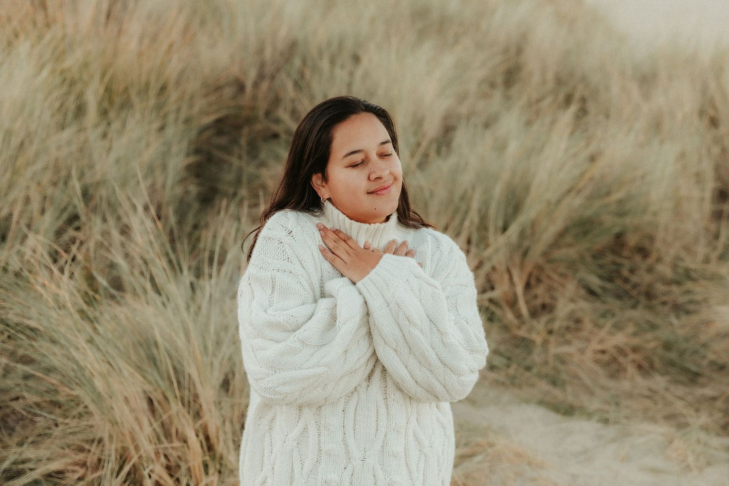 A figure in a cozy white sweater poses expressively against a backdrop of dried grass in warm natural lighting.