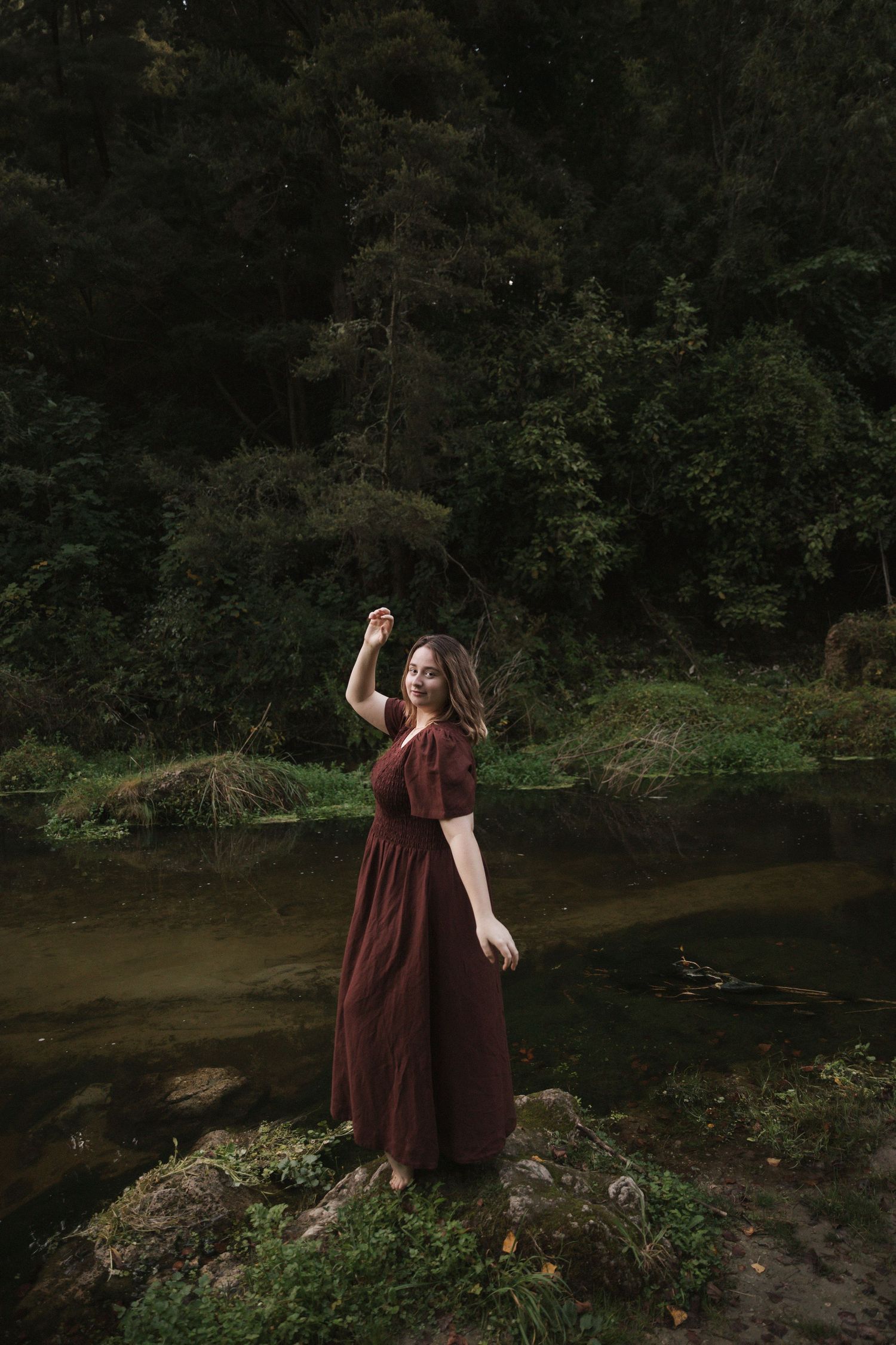 A whimsical scene by a forest stream with a burgundy dress and natural surroundings.