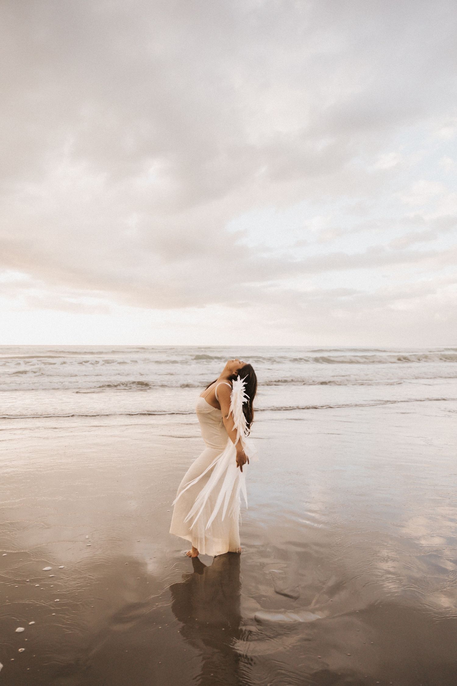 A person in a flowing white dress stands at the edge of the ocean during sunset, creating a dreamy coastal scene.