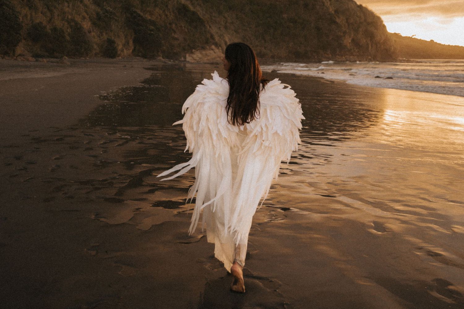 A person in a flowing white dress walks along a sandy beach at sunset with golden light reflecting on the water.