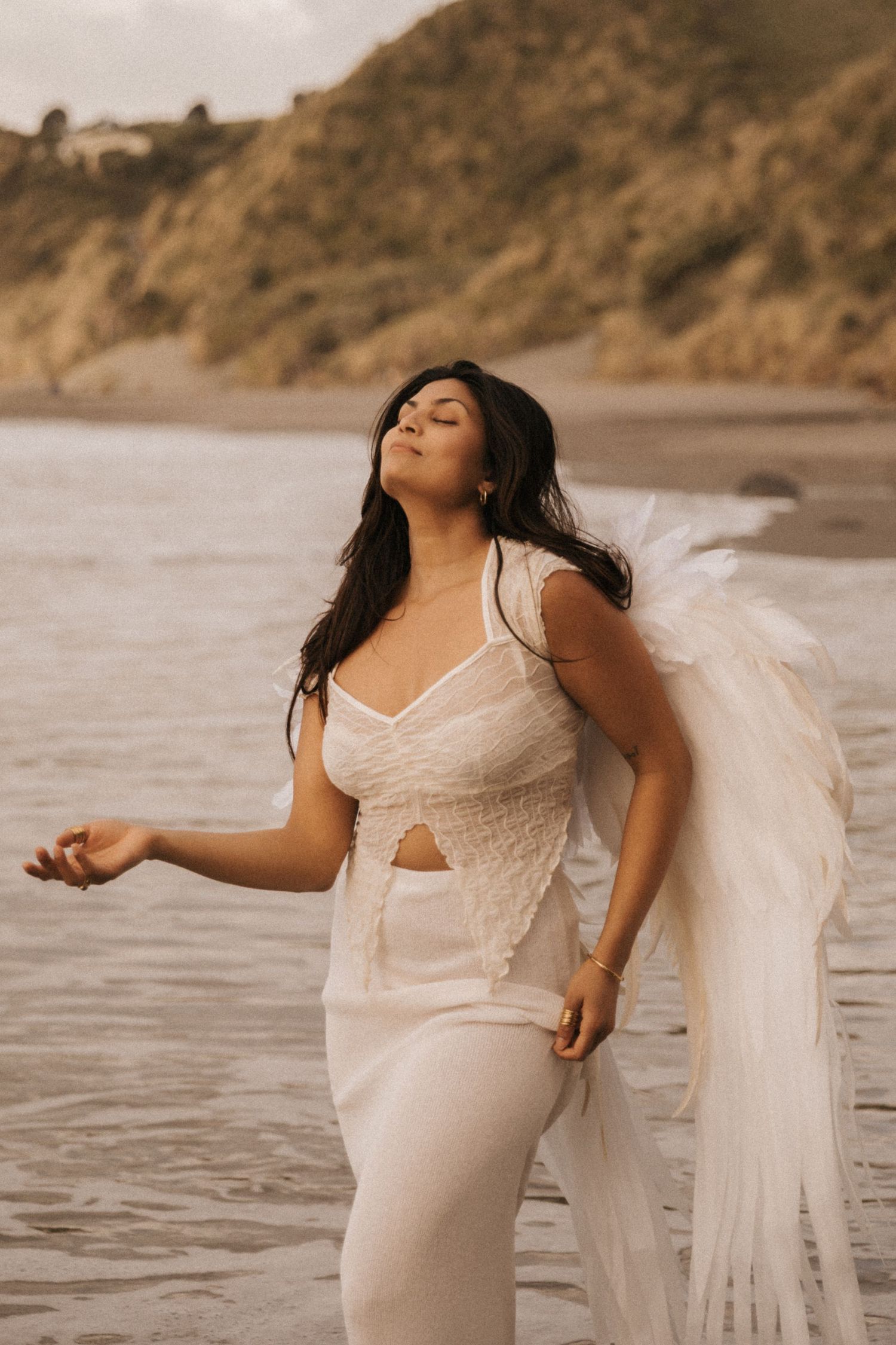 A person in a flowing white dress poses expressively on a beach at sunset with cliffs in the background.