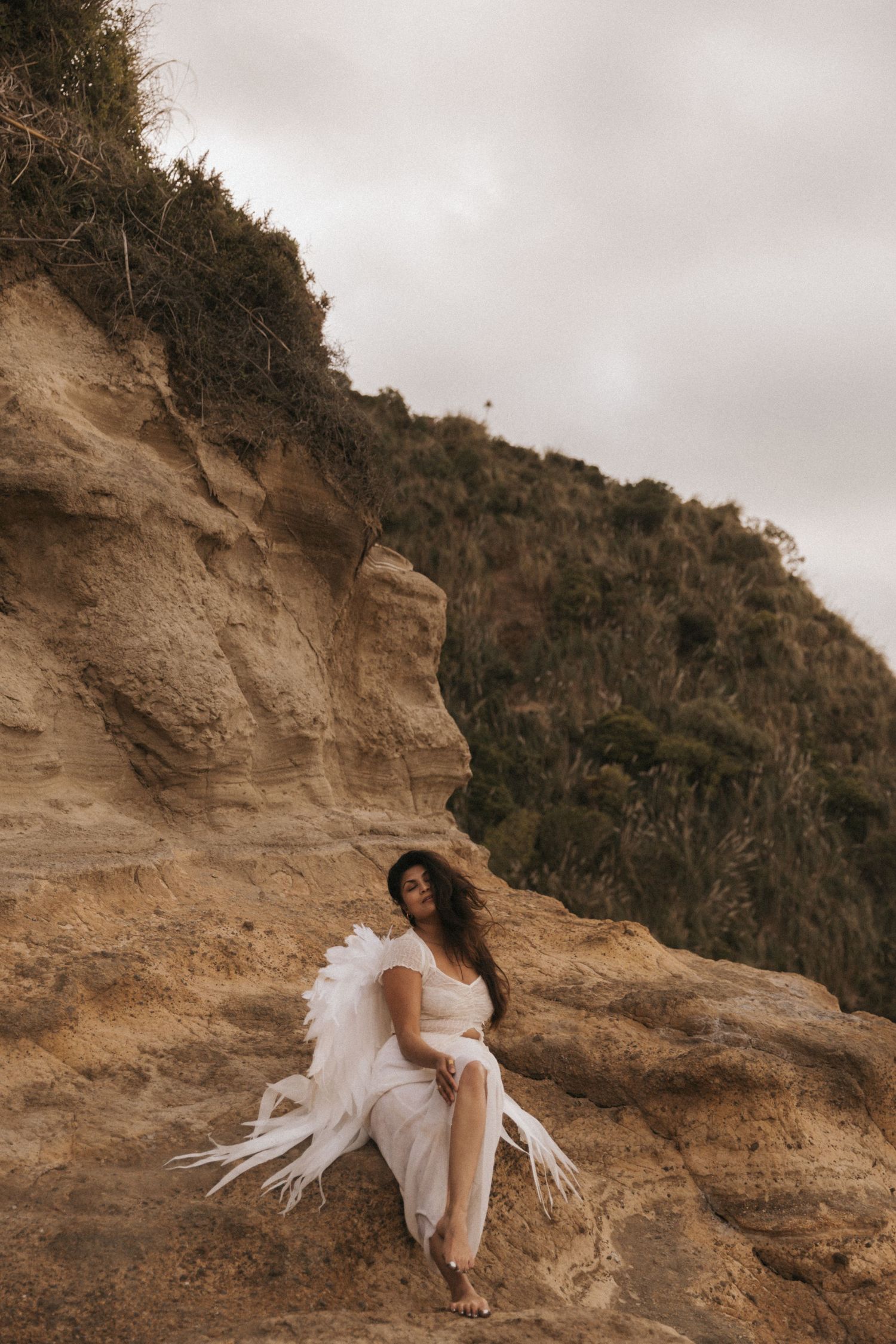 A figure in a flowing white dress poses gracefully against rugged coastal cliffs under an overcast sky.