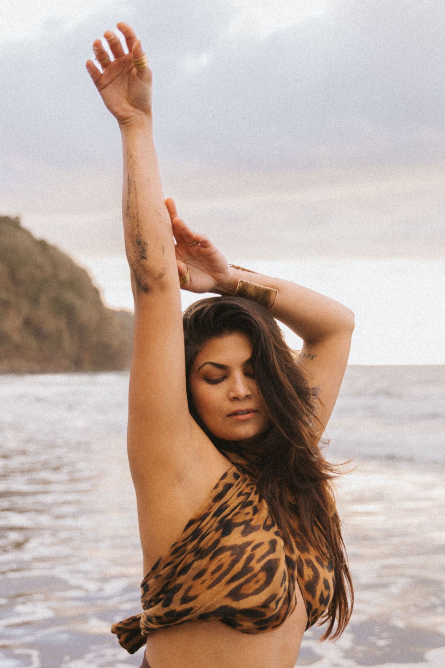 Woman in leopard print swimsuit poses with arms raised on beach at sunset.