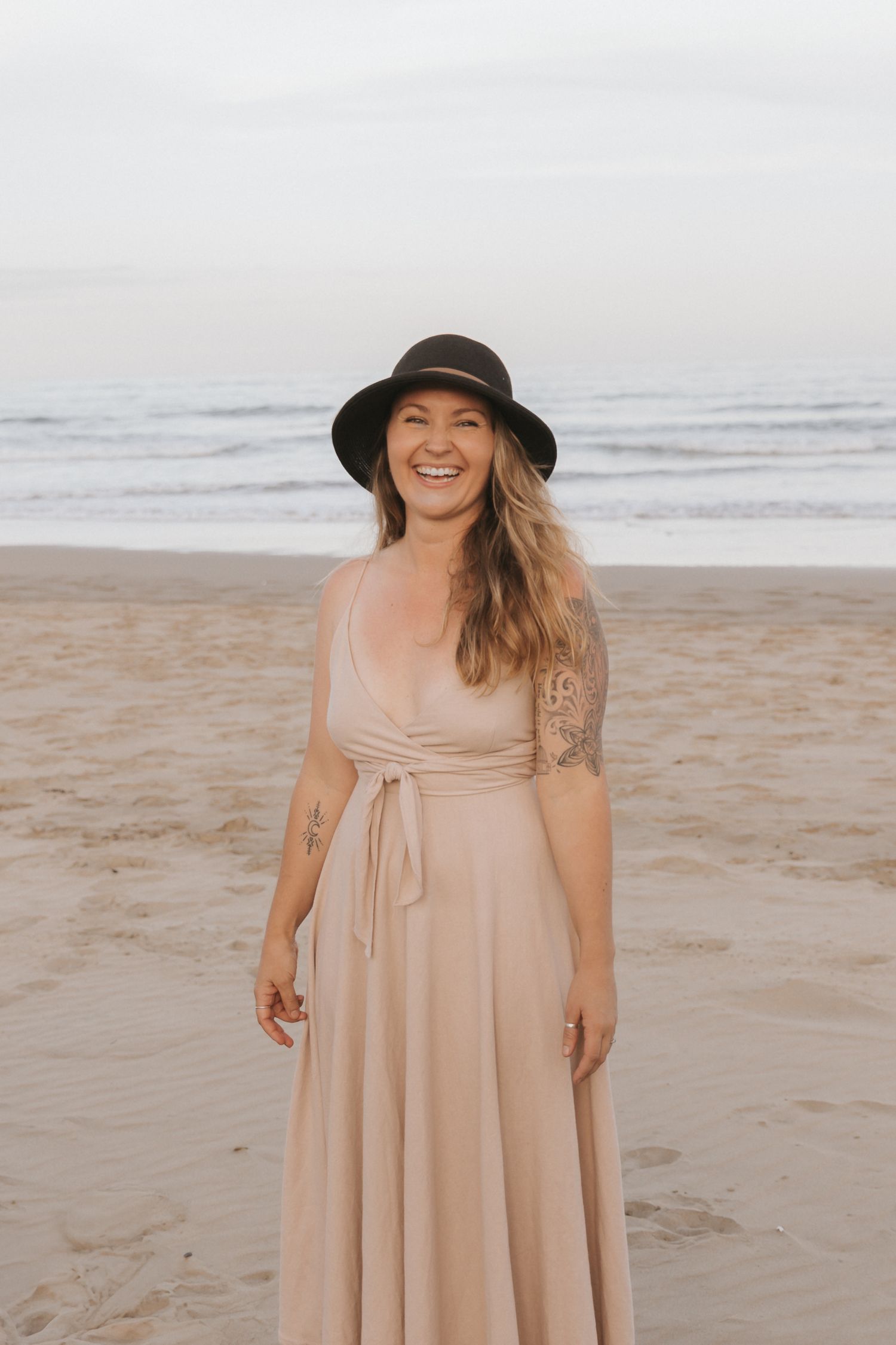 A person in a flowing beige dress and black sun hat stands on a sandy beach with ocean waves in the background.