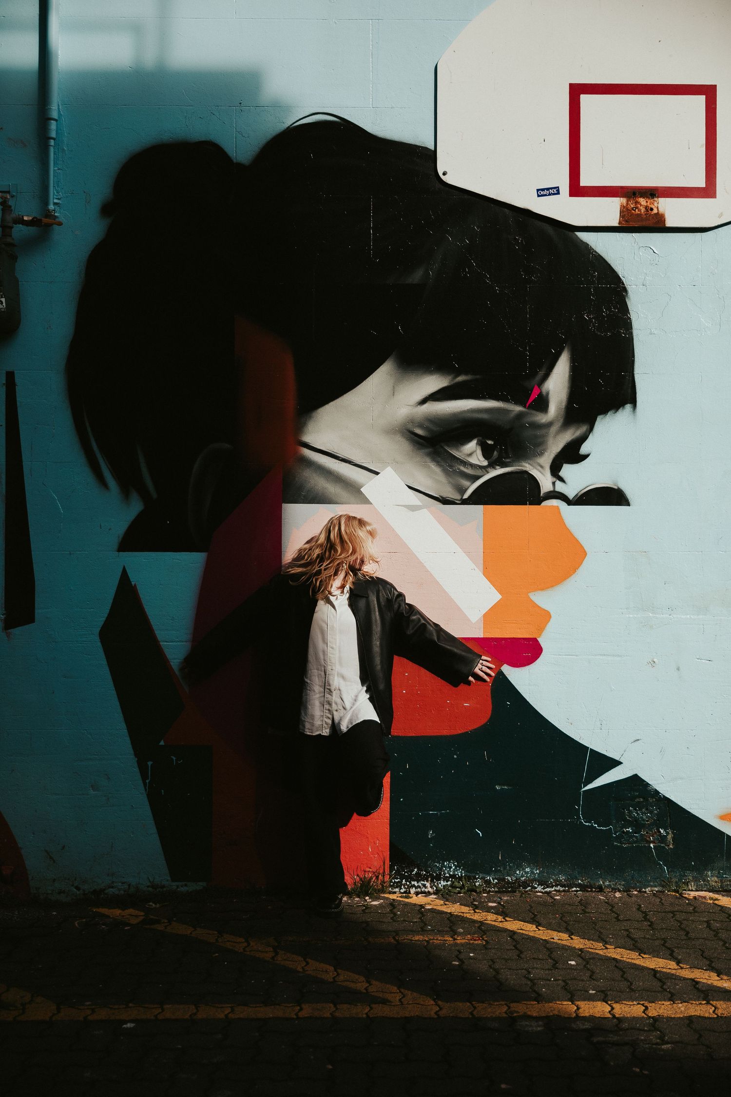 Person in white shirt stands against colorful street art mural featuring abstract portrait on turquoise wall with basketball hoop.