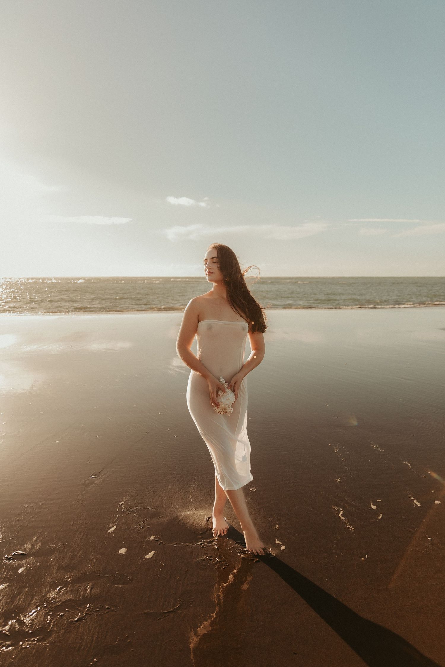 Expectant mother in white dress poses on reflective beach at sunset.