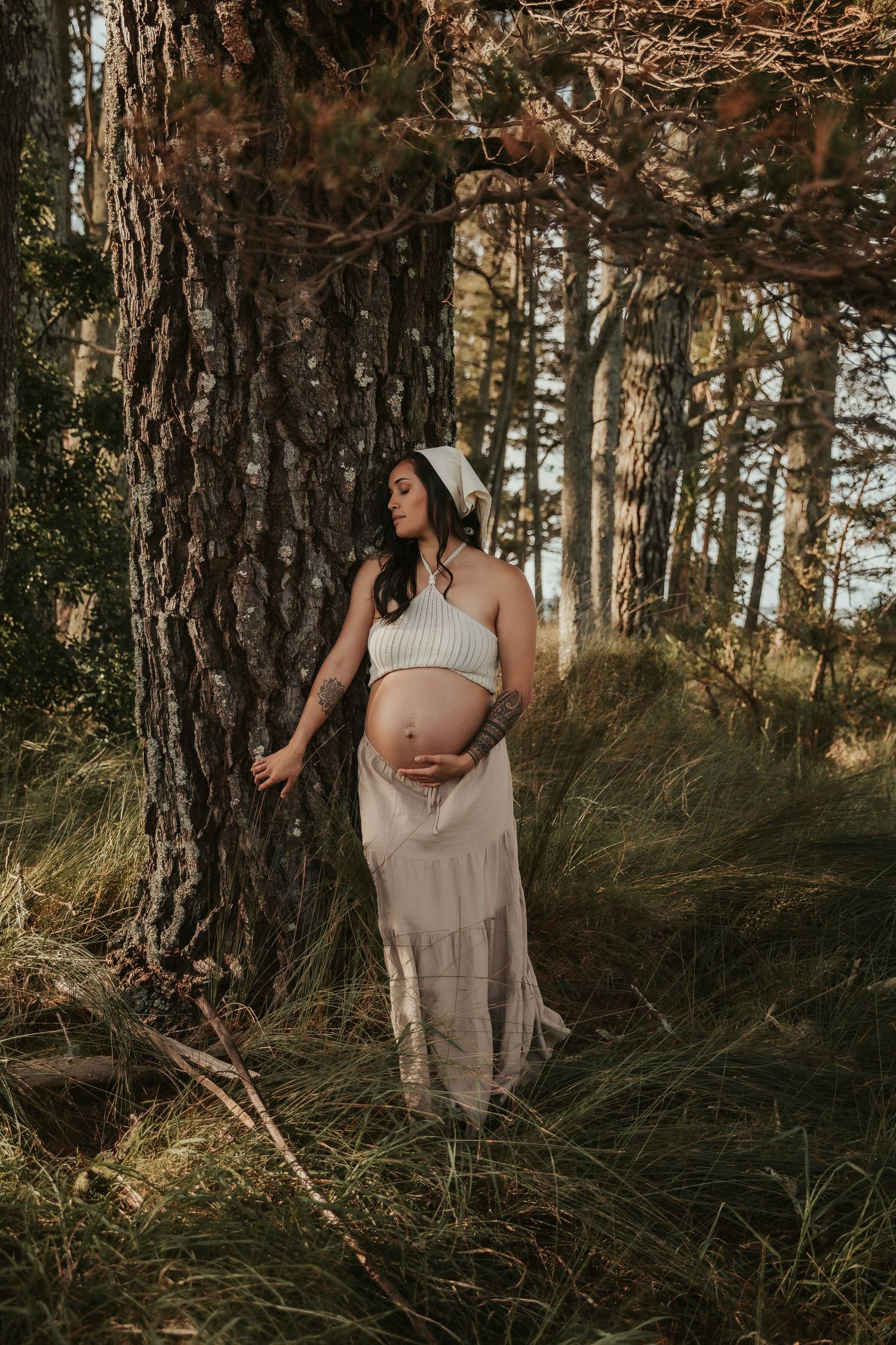 Maternity photoshoot in a serene forest setting with warm golden light filtering through tall pine trees.
