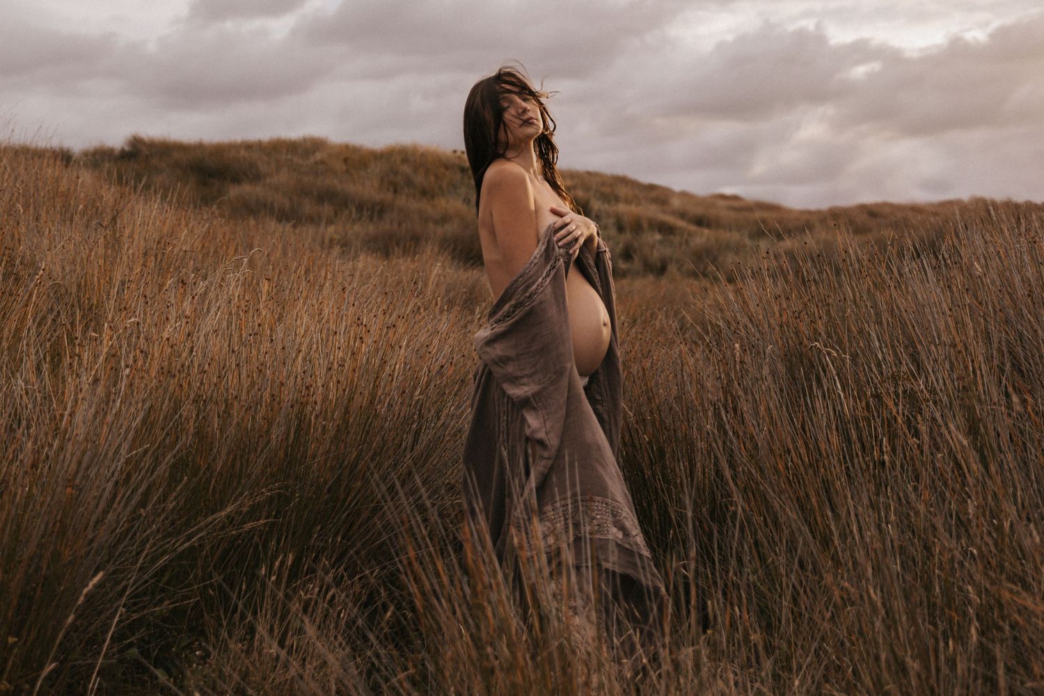 Artistic maternity photos taken in a rustic field setting with dramatic cloudy sky.