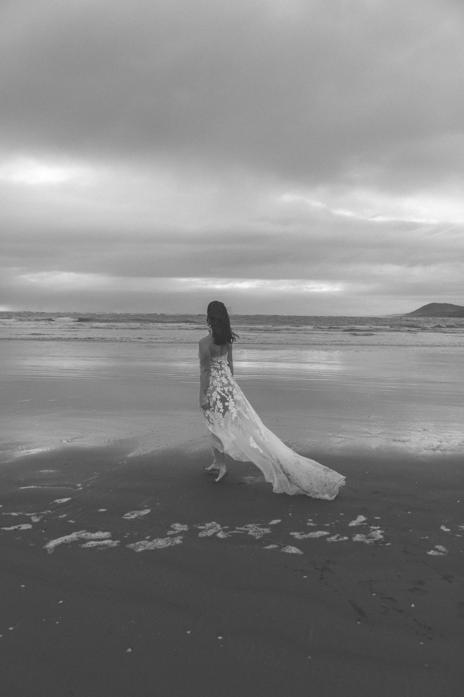 Dramatic black and white photo of bride walking on empty beach with mountains in distance.