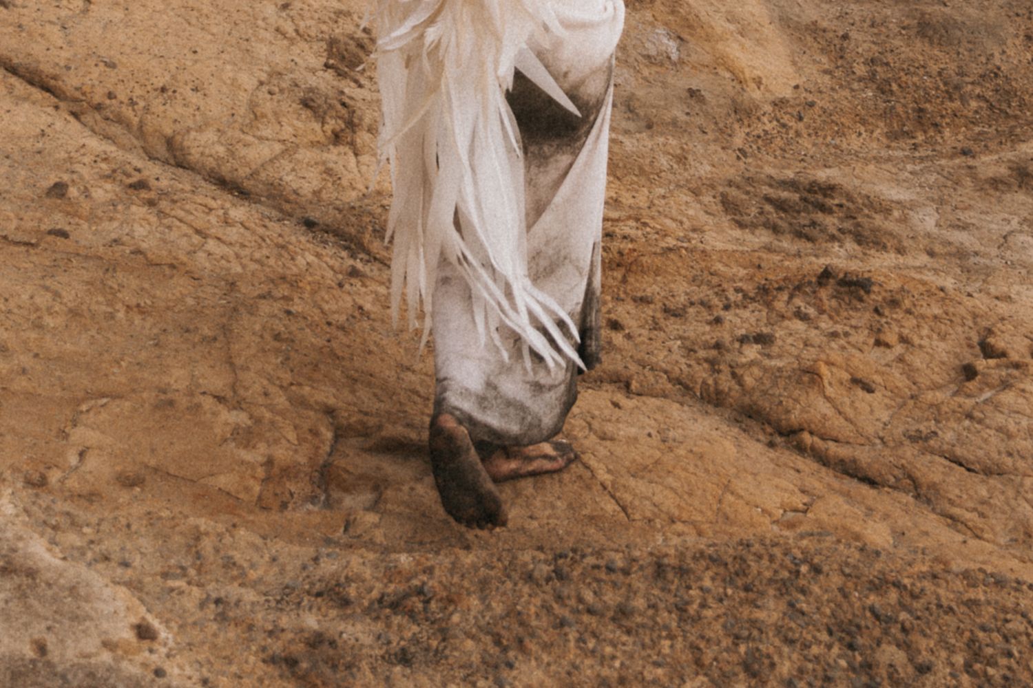 A person wearing flowing white fabric walks along a dusty brown earthen surface in an artistic composition.